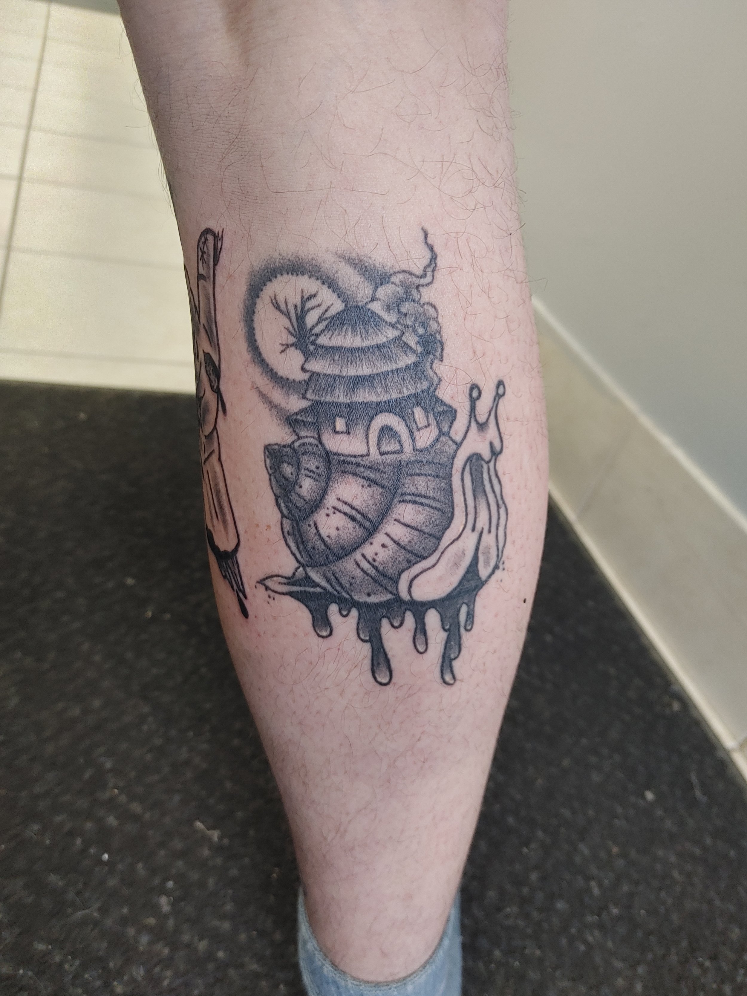 Tattoo of a bug with a house on its back, surrounded by a circular pattern, on person's lower leg.