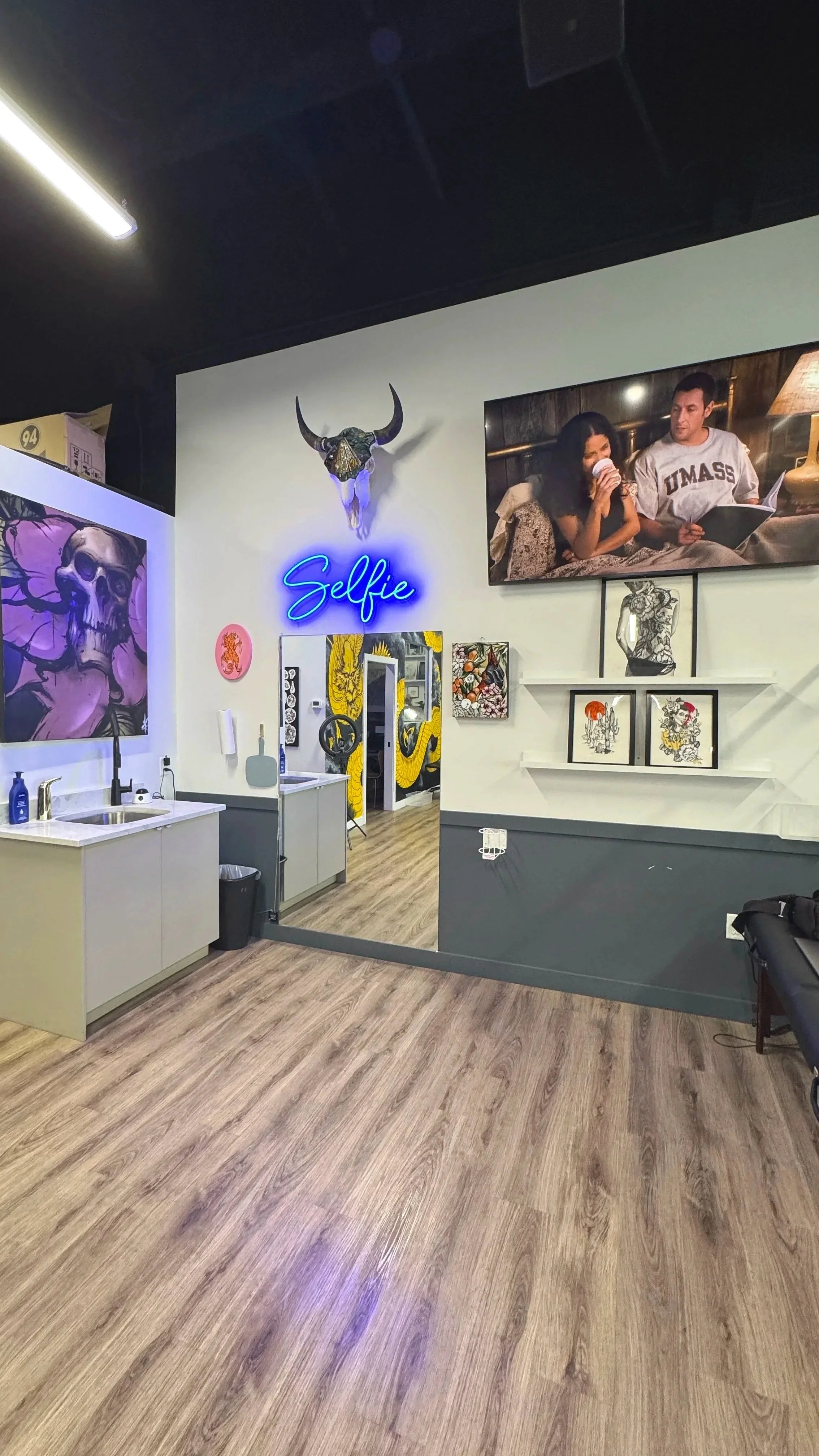Interior of a tattoo or art studio with a neon blue sign that says 'Selfie' on the wall, framed artwork, a mounted bull skull, a large LED screen showing a couple, and a mirror reflecting more of the decorated space.