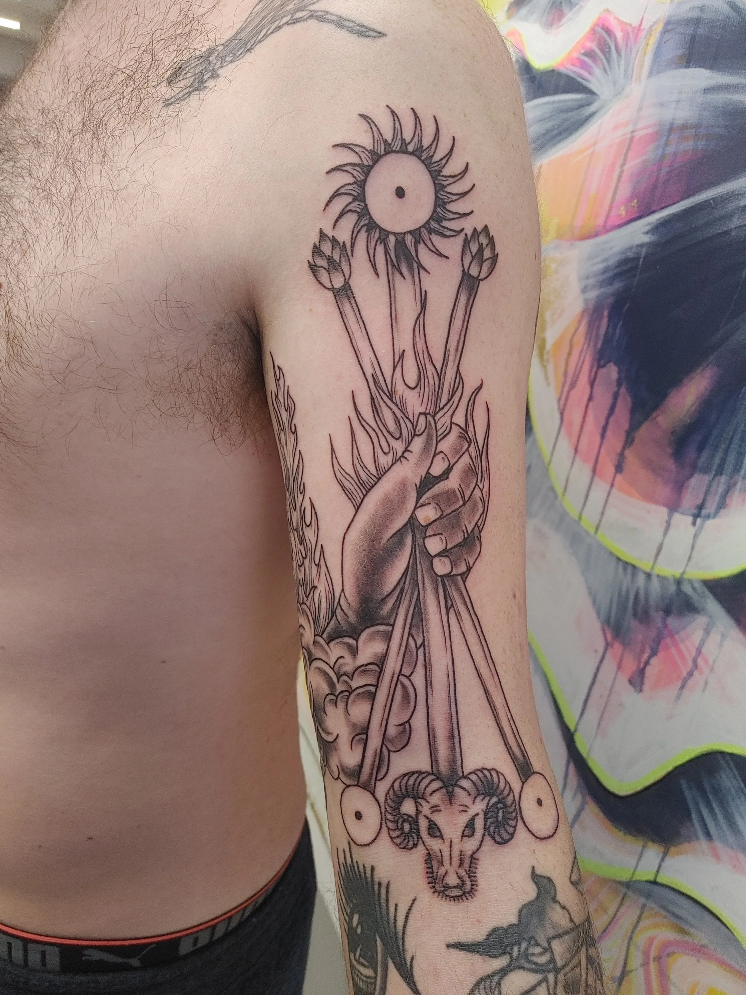 A tattoo on a person's arm featuring a hand holding a staff with a bow and arrow, a sun with a face, a lion's face, and clouds, with surrounding flames and decorative elements.