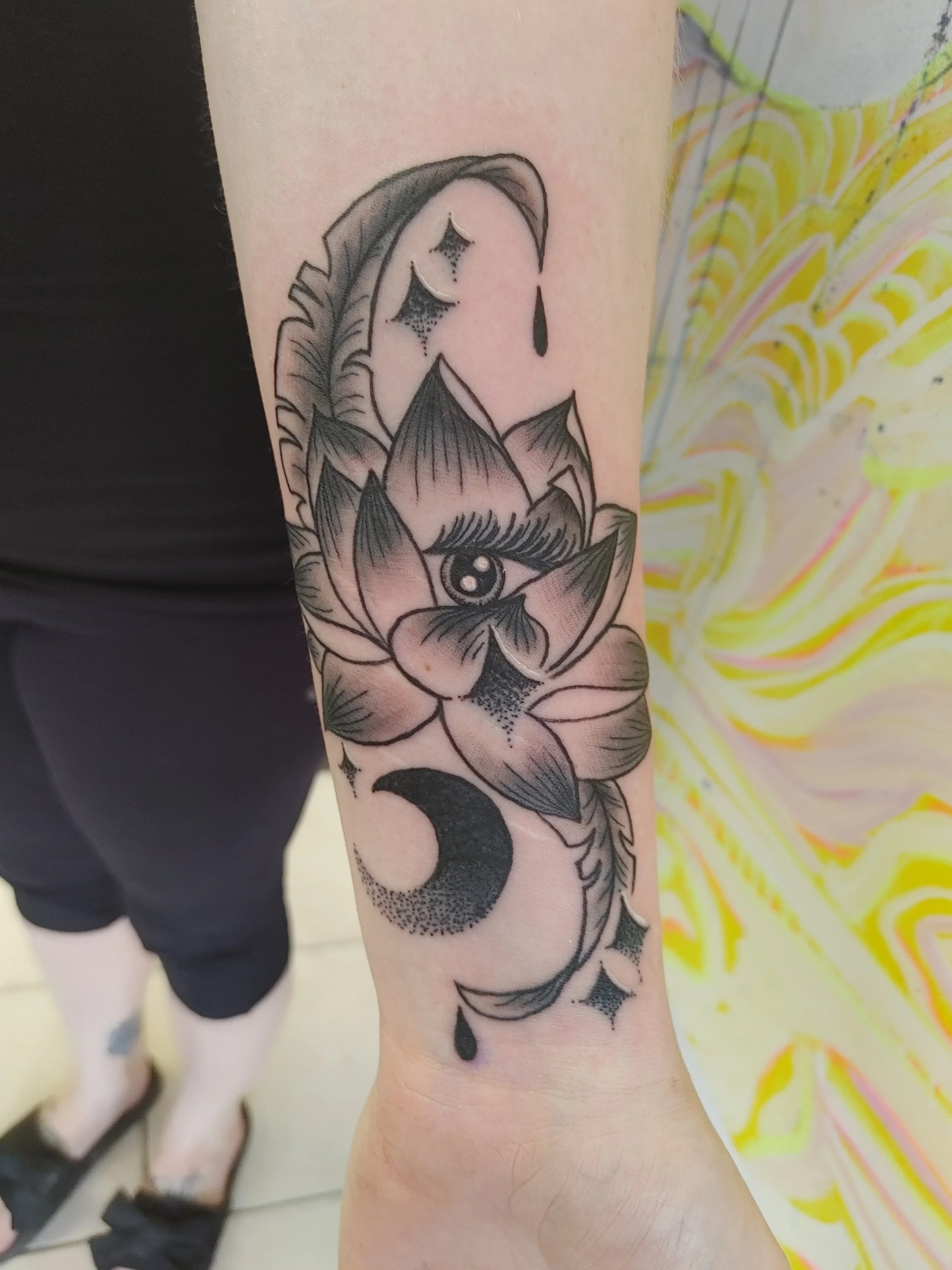 Black tattoo on arm featuring a lotus flower with an eye in the center, surrounded by abstract designs and dark watercolor shading.