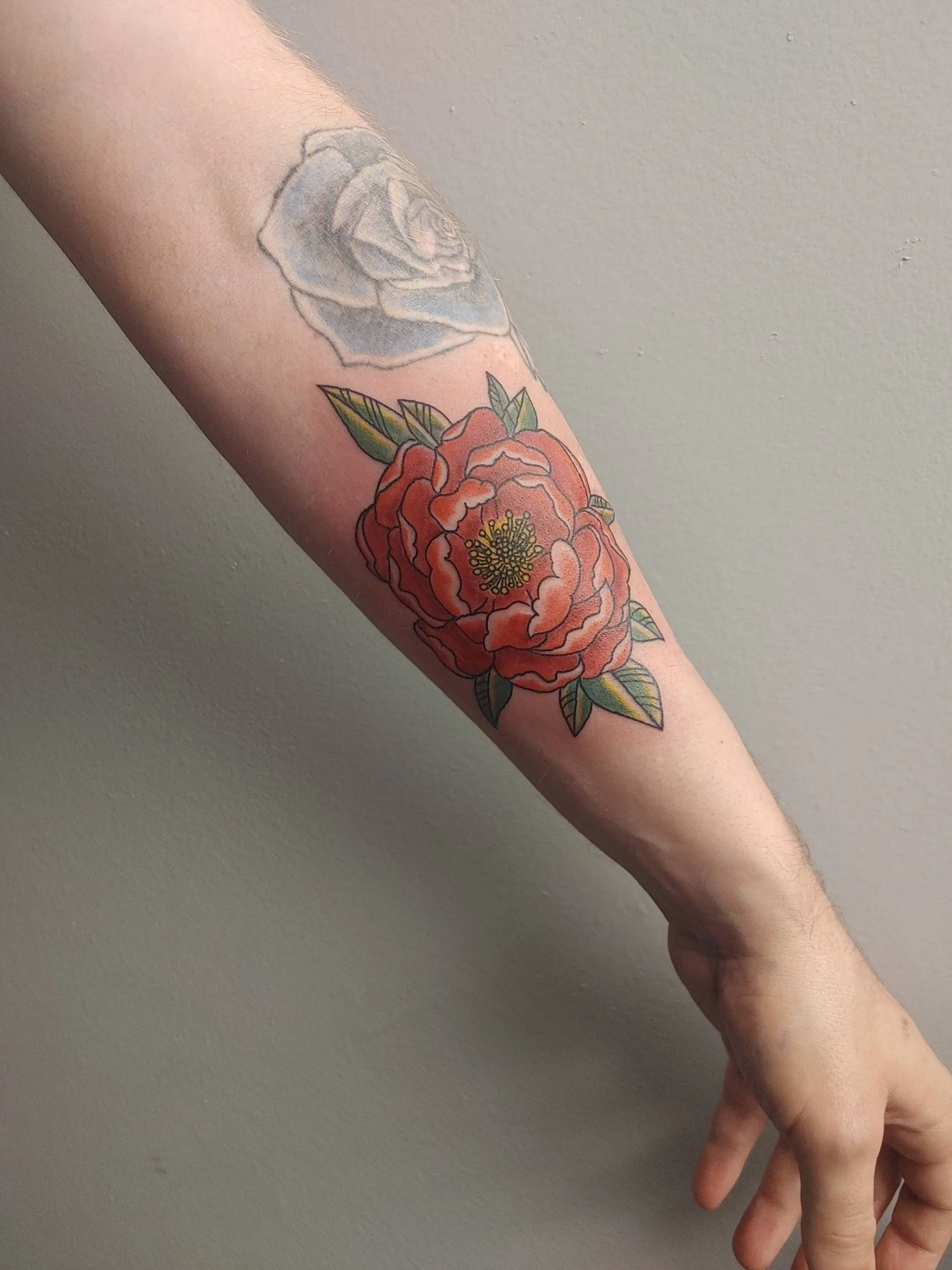 A person's arm with two tattoos, one above the other. The upper tattoo is a shaded gray and black rose. The lower tattoo is a colorful, detailed red and orange flower with green leaves.