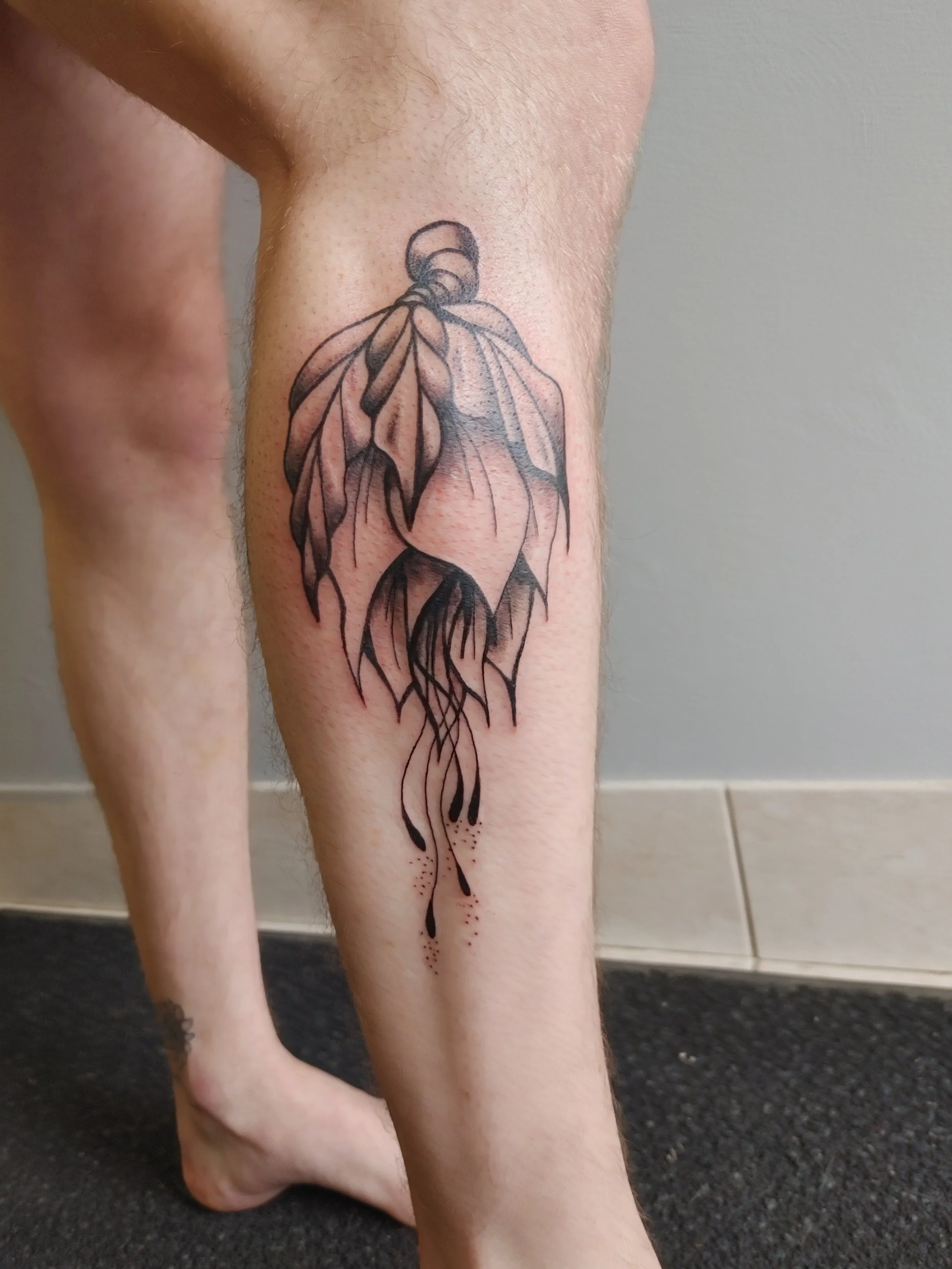 A black ink tattoo of a stylized plant with elongated leaves and flowing roots on the person's lower leg.