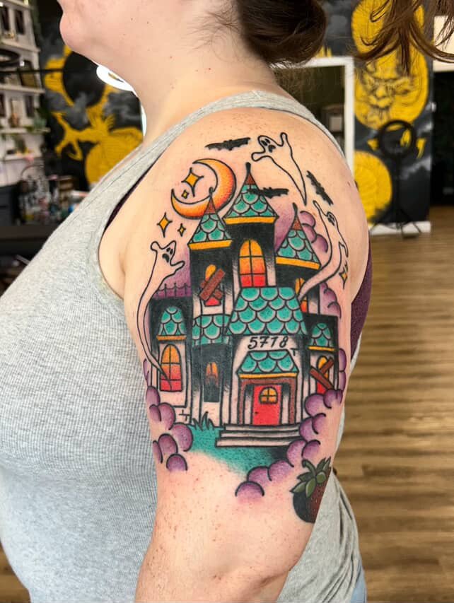 Colorful tattoo of a haunted house with bats, ghosts, and a crescent moon with stars on a person's upper arm and shoulder.