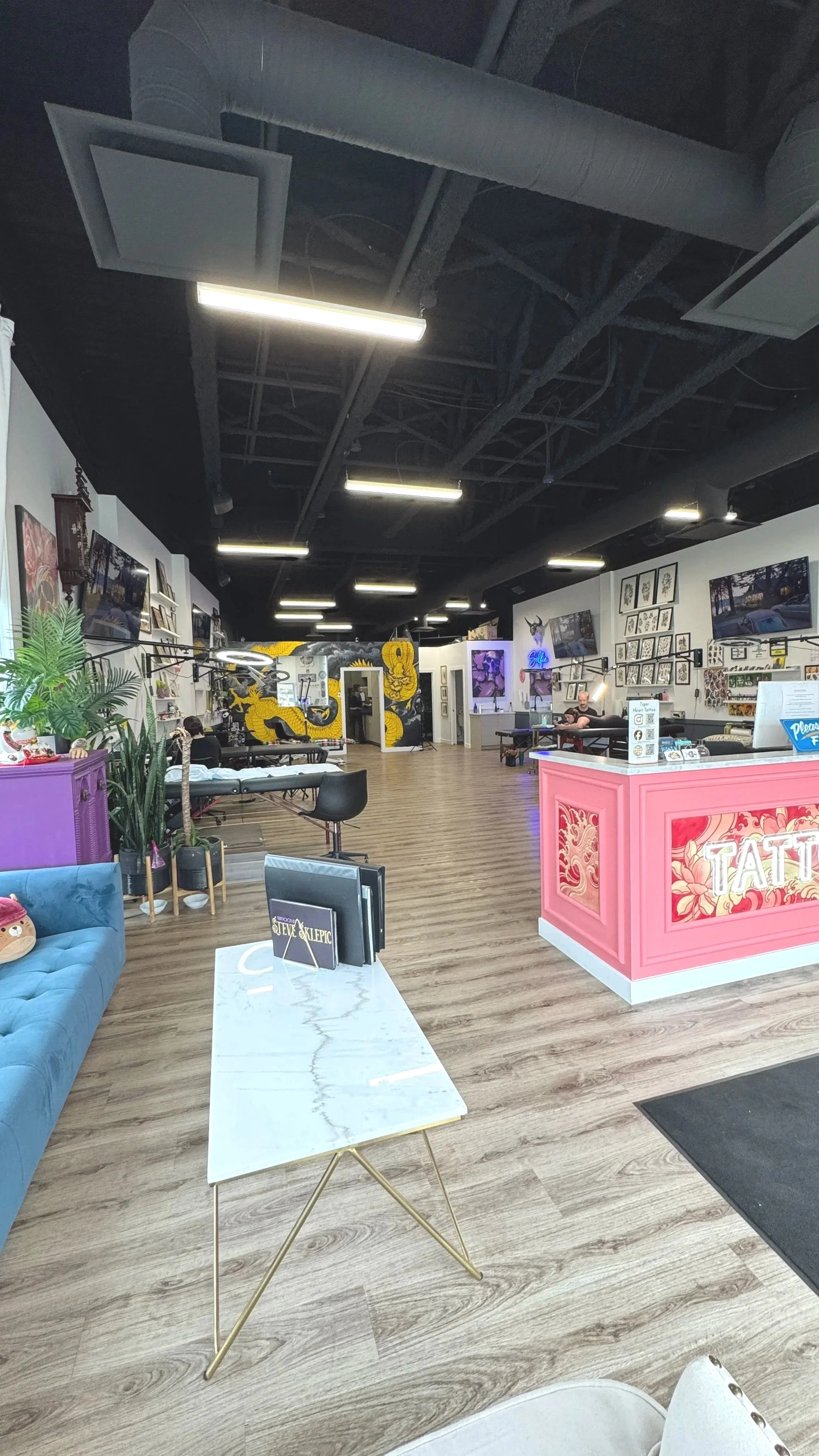 Interior of a tattoo studio with black ceiling, wood flooring, various wall decor, display screens, and a pink reception desk.