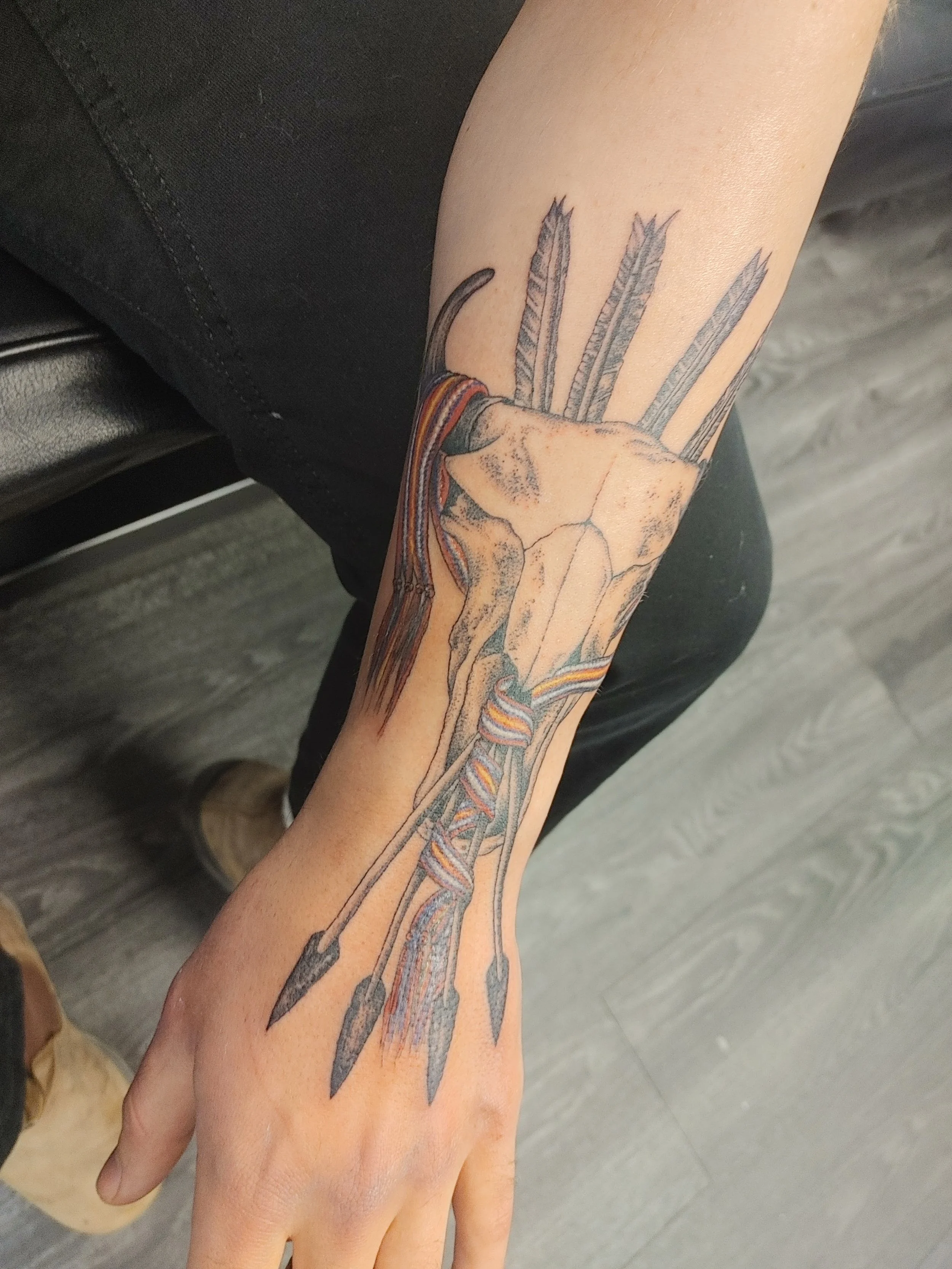 Tattoo of a deer skull with antlers on a person's forearm, with feathers, tribal designs, and colorful ribbons.