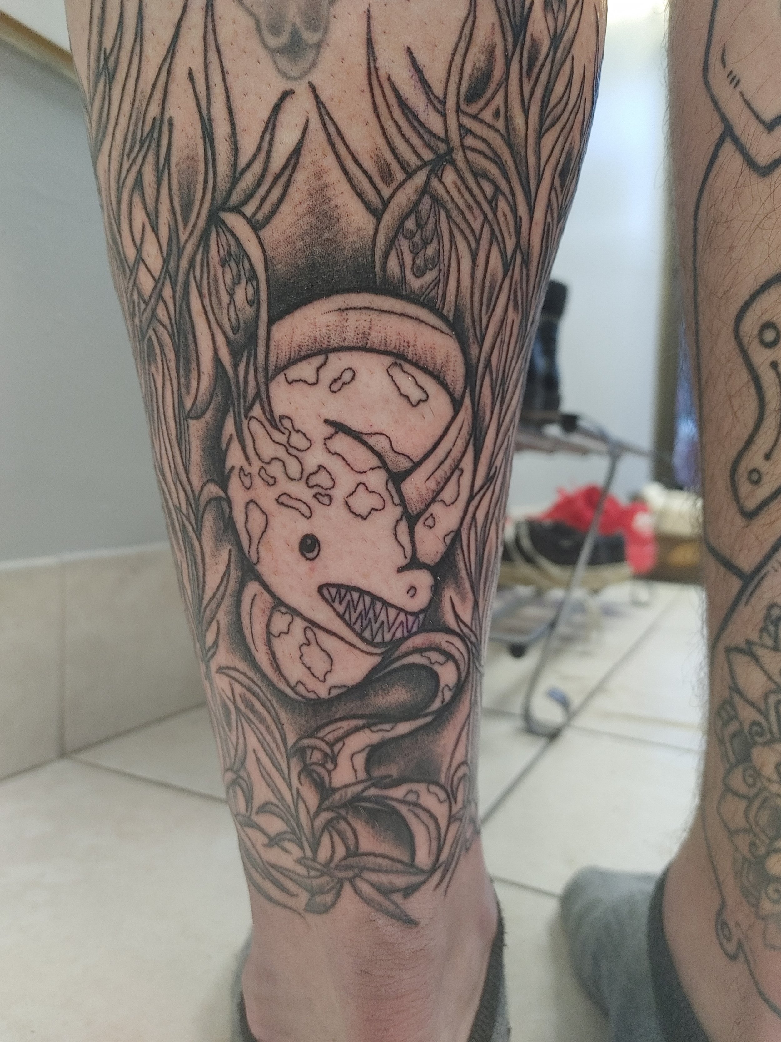 A tattoo of a cartoonish dinosaur head on a person's lower leg, with sharp teeth and a playful expression, surrounded by abstract line designs.
