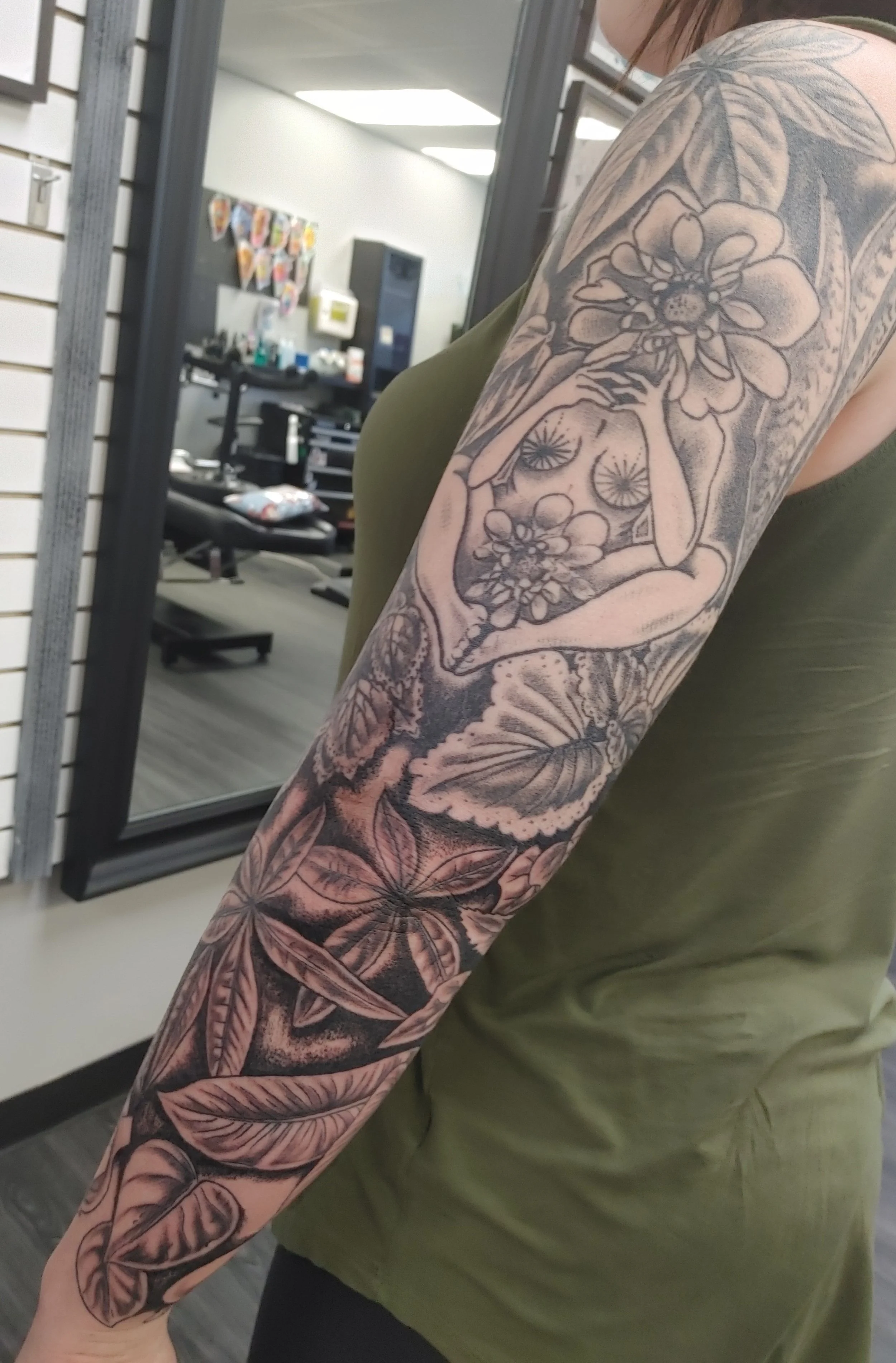 Close-up of a person's arm with a large, detailed black and gray tattoo featuring flowers, leaves, and abstract designs, taken in a tattoo parlor.