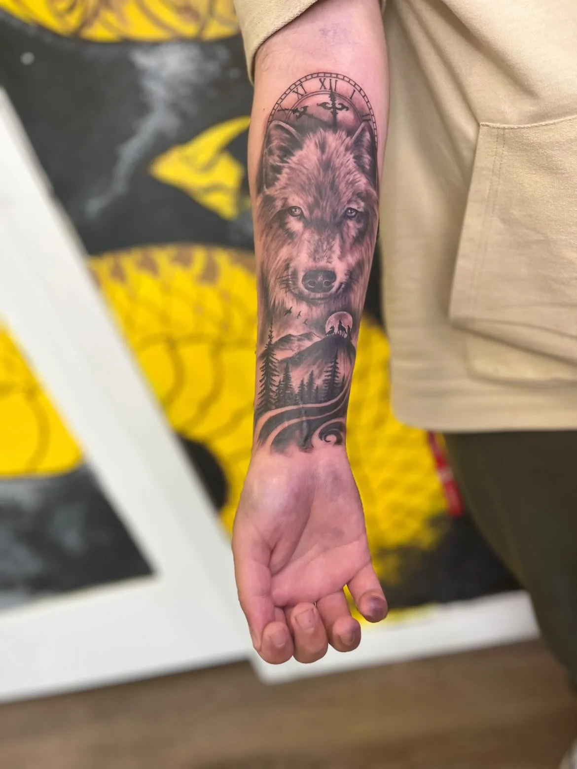 A tattoo on a person's forearm featuring a wolf's face, a clock with Roman numerals, mountains, pine trees, a full moon, and birds in the sky.