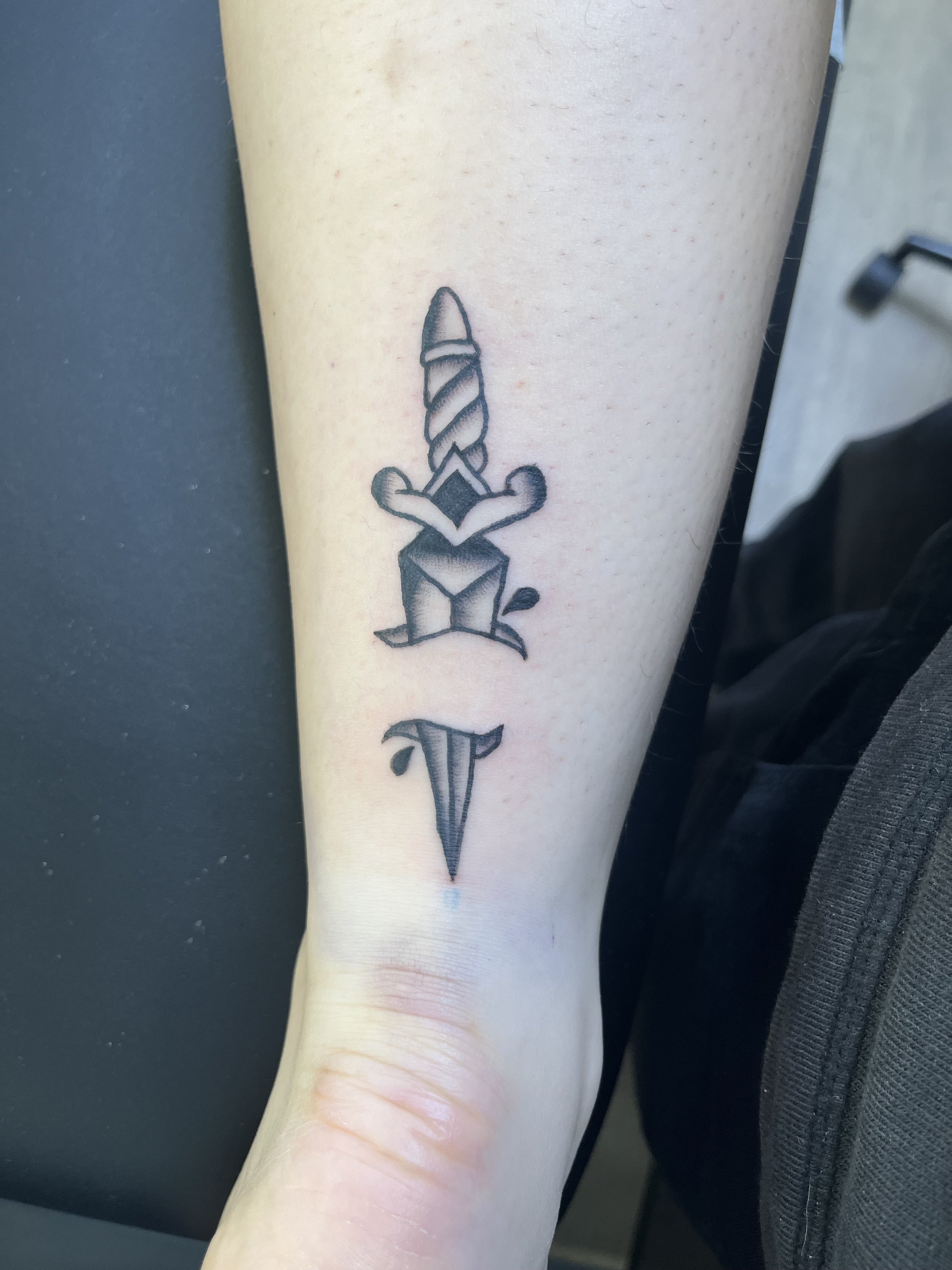 A tattoo of a sword with a diamond-shaped guard on a person's inner forearm.