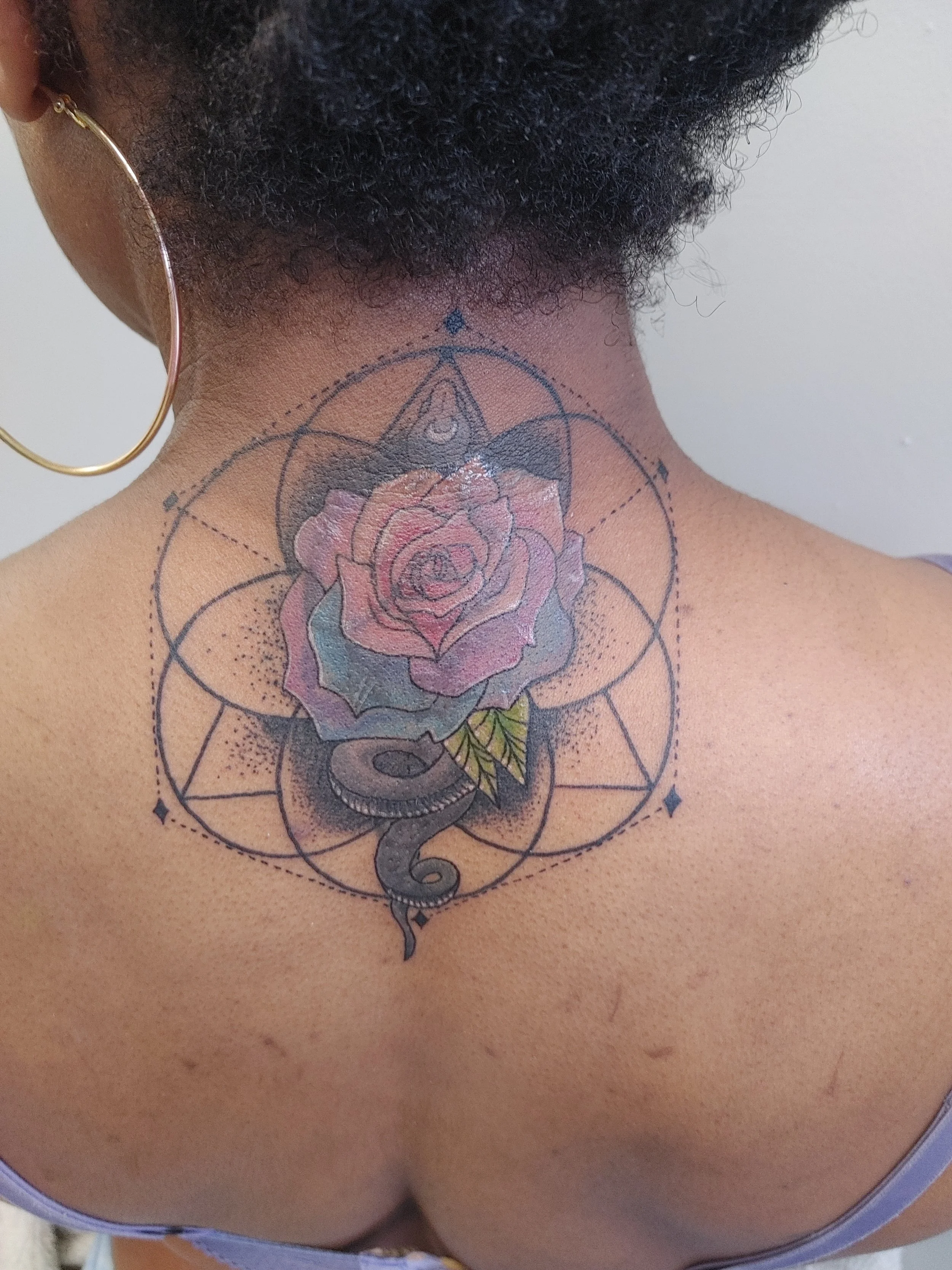 Close-up of a person's neck and upper chest, showing a detailed colorful tattoo of a rose, geometric shapes, and a snake, with the person's head slightly tilted and wearing a gold hoop earring.
