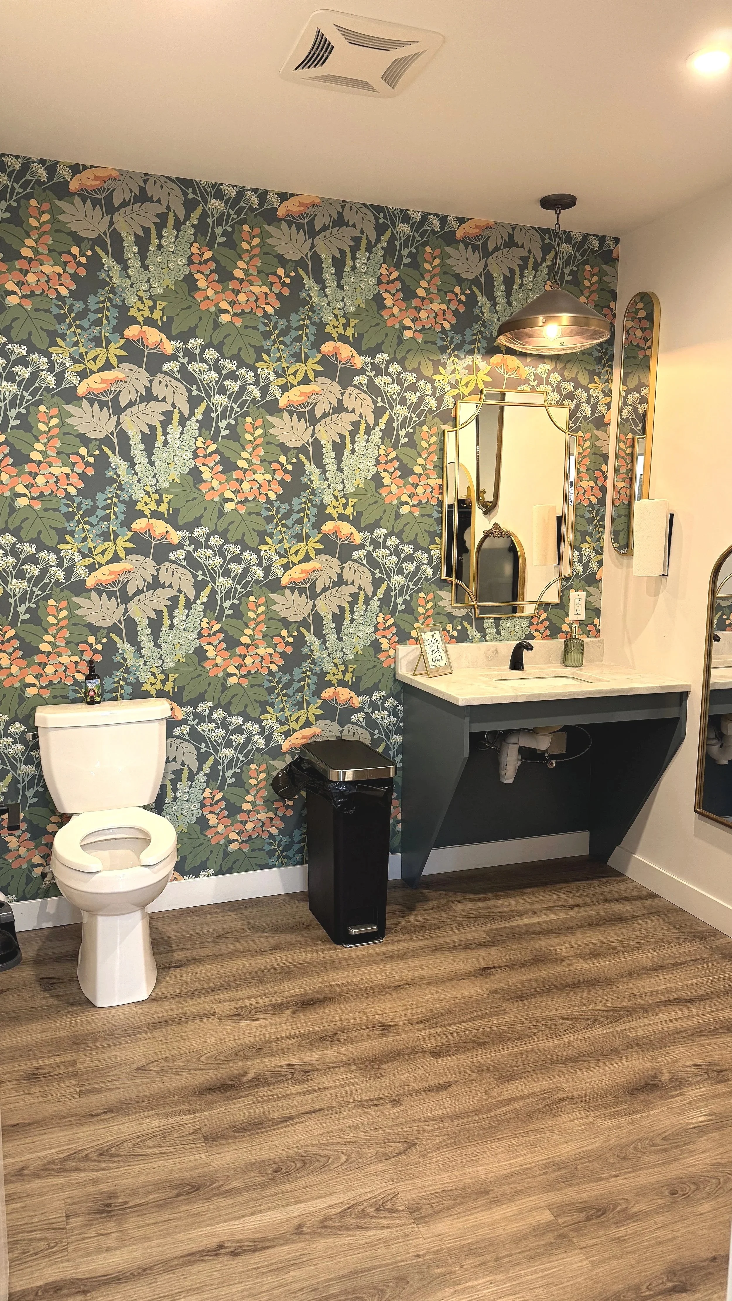 Restroom with tropical floral wallpaper, a white toilet, a black trash bin, and a sink with a gold-framed mirror and gold fixtures.
