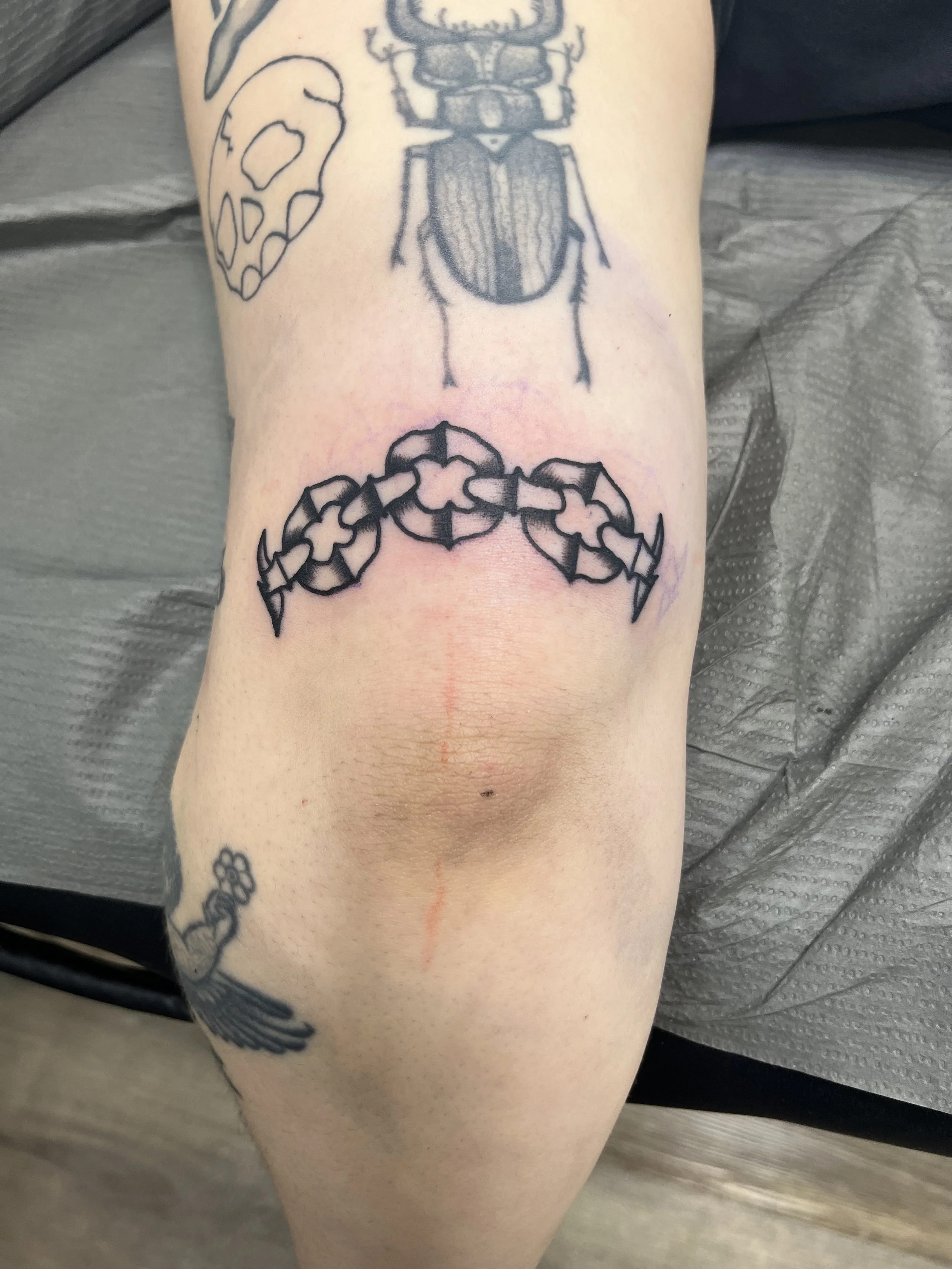 Tattooed arm with various black ink tattoos, including a skull outline, a detailed beetle, and a chain-like design, resting on a gray surface.