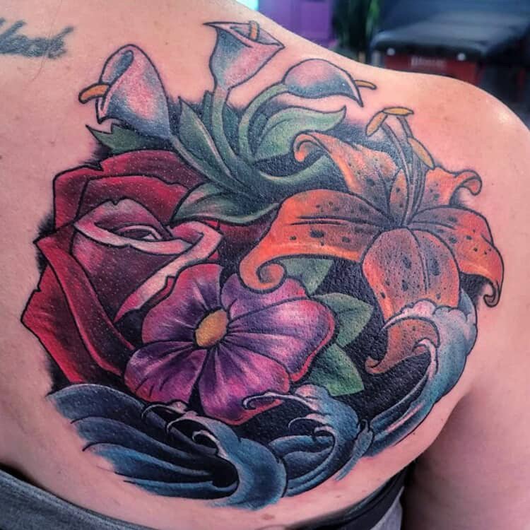 Colorful tattoo of a bouquet of flowers on shoulder, including a red rose, a purple flower, and an orange lily with green leaves and stylized black and blue waves in the background.