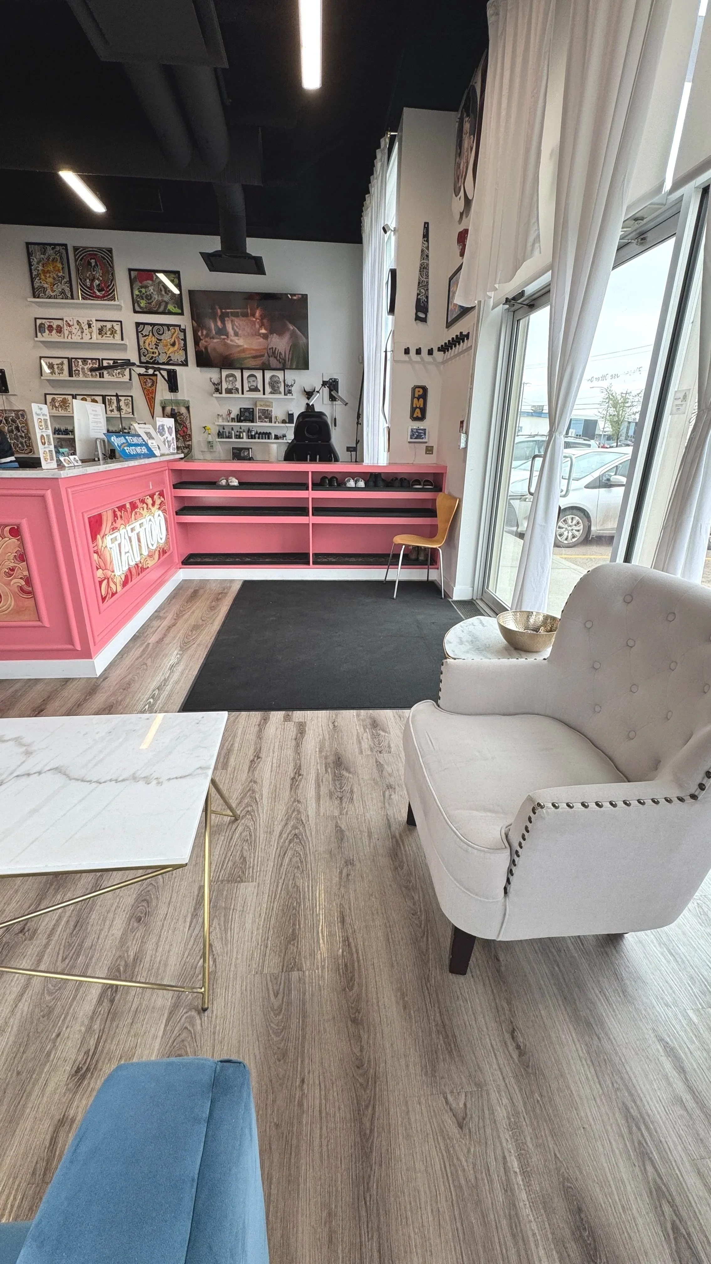 Interior of a tattoo shop with a pink counter, artwork on the walls, a TV, a chair near the window, a white armchair, and a marble coffee table.