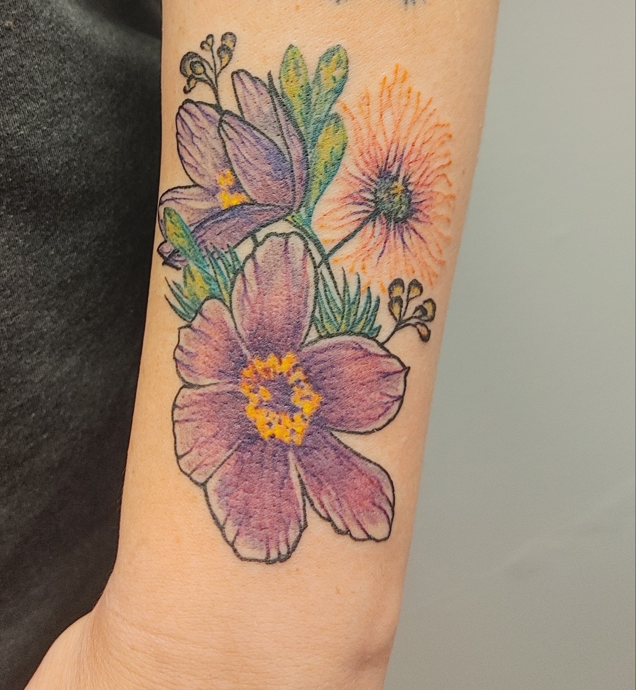 Colorful tattoo of purple and pink hibiscus flower with yellow center, set on the person's arm with green leaves and orange accents.