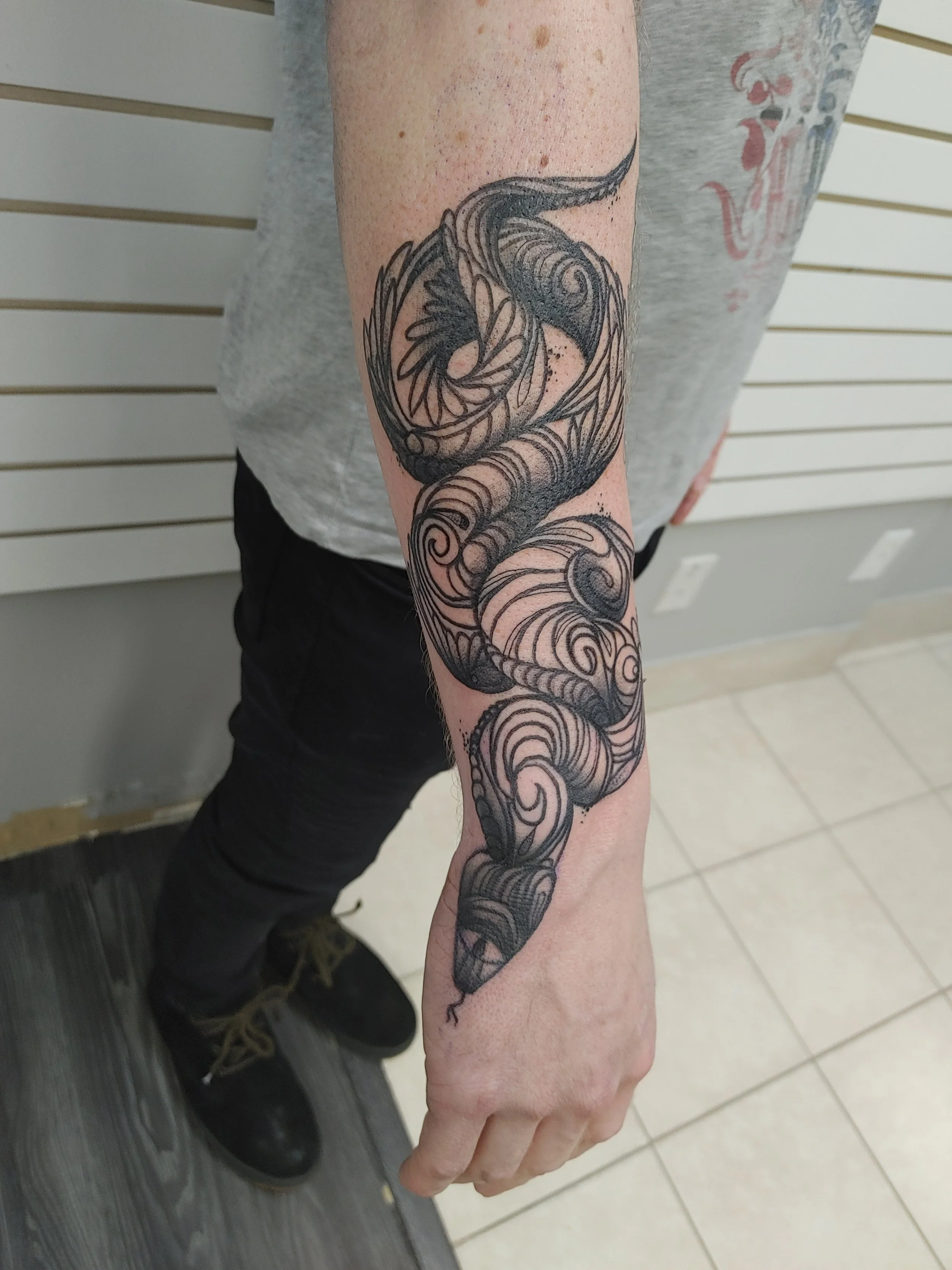 A person's forearm with a detailed black and gray tattoo of a stylized seahorse surrounded by swirling patterns.