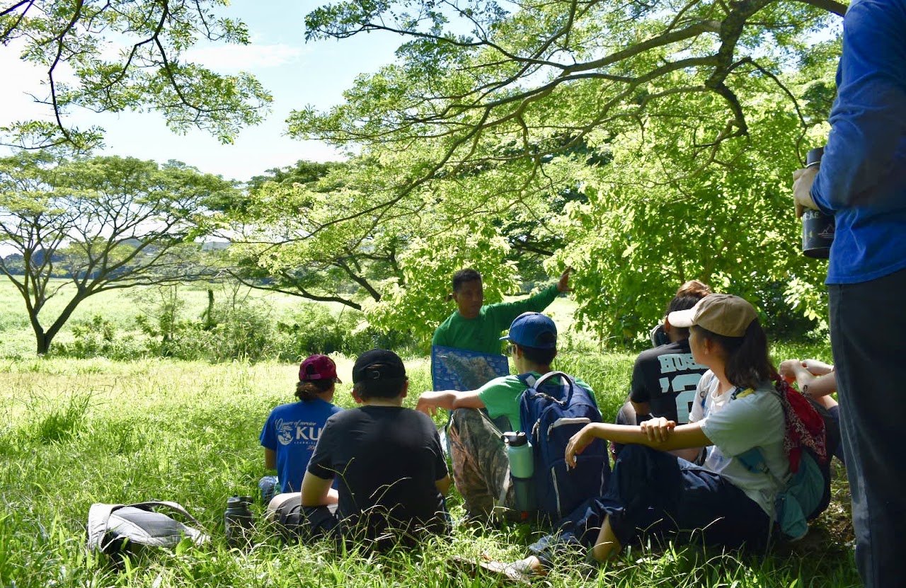 Teacher (kumu) holding a map of O'ahu and pointing towards a group of students gathered in an outdoor pasture for a lesson.