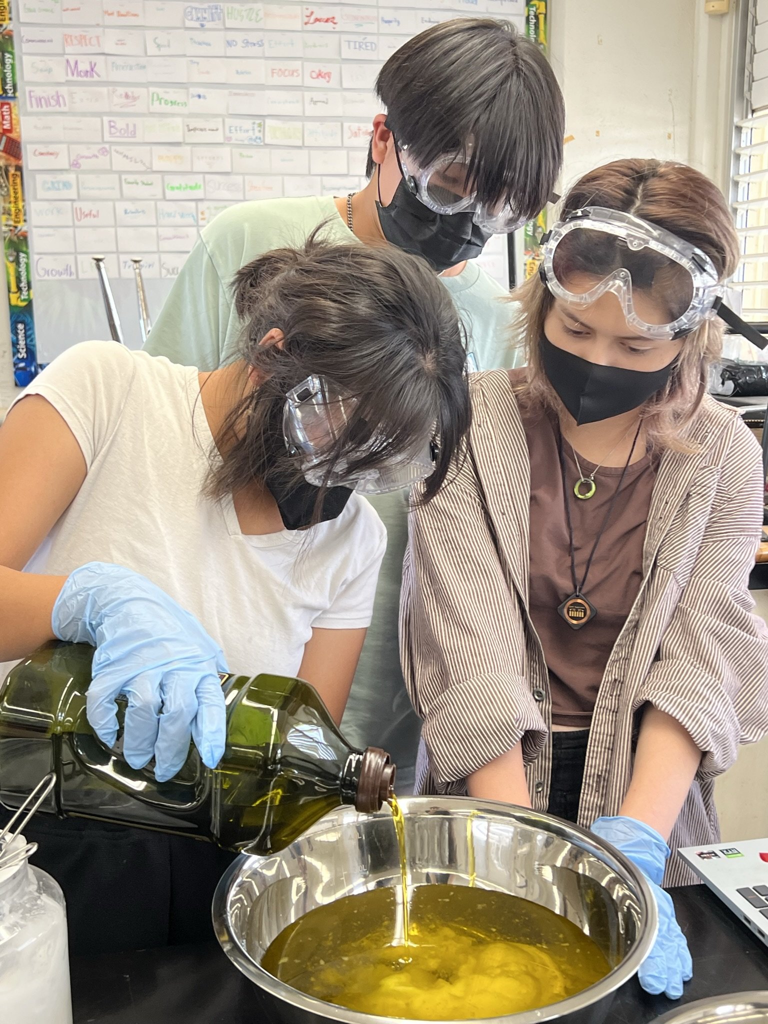Three young adults wearing safety goggles in a lab room and pouring oil into a bowl.