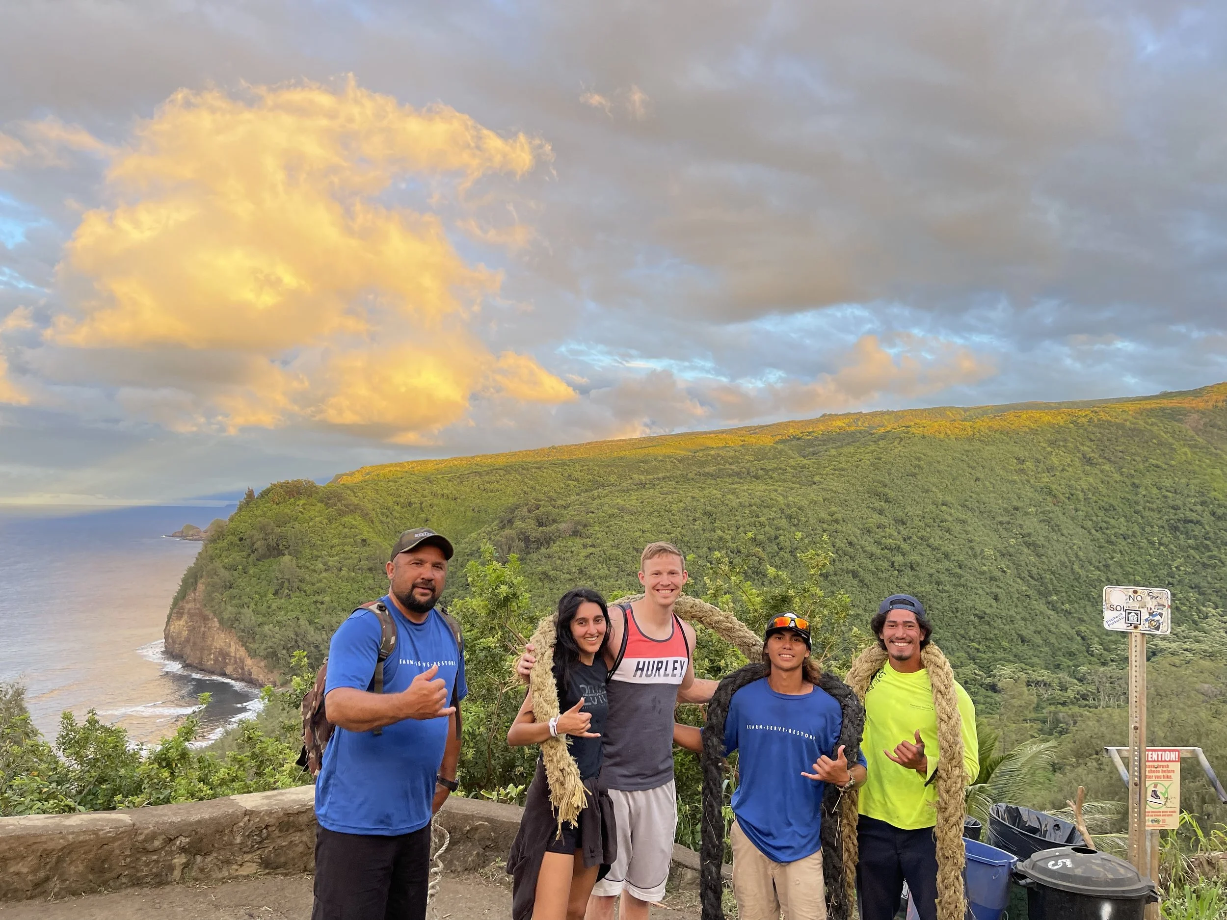 Five cheerful Kupu participants at the peak of Polulu Trail, smiling and making shaka gestures, while carrying washed-up rope.