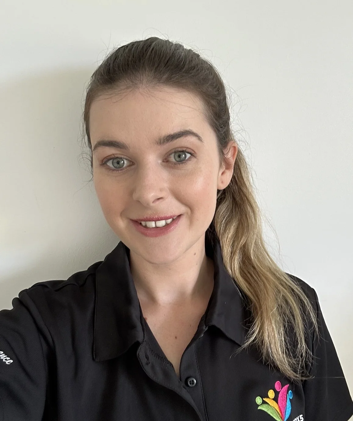 Female staff member Emma. Emma has long straight brown hair in a ponytail, she is wearing a black polo uniform shirt and she is smiling.