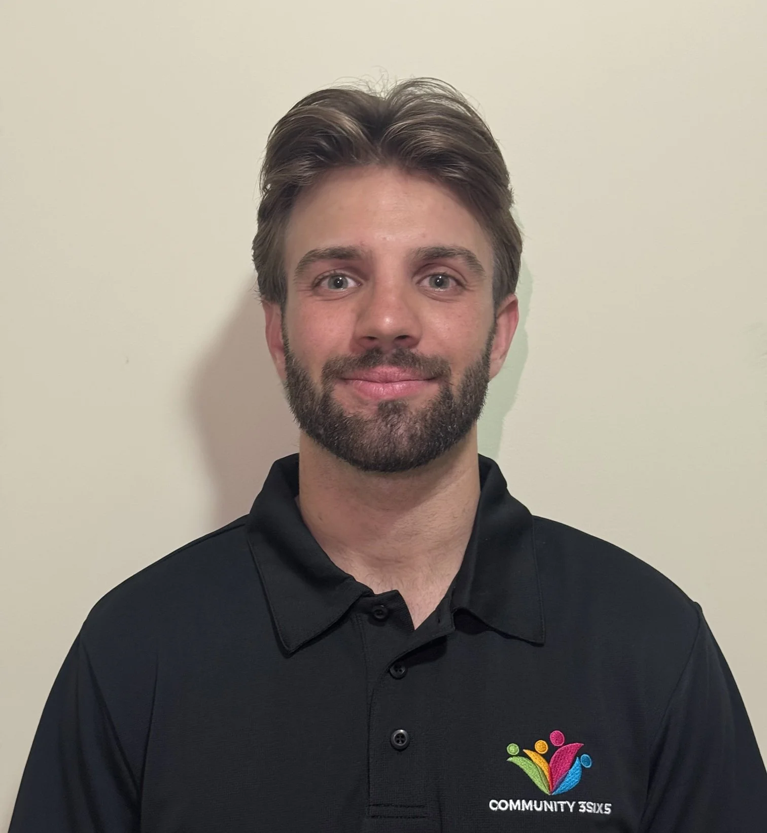 Male staff member Zander. He has short brown hair and a short brown beard. He is wearing a black uniform polo and he is smiling.