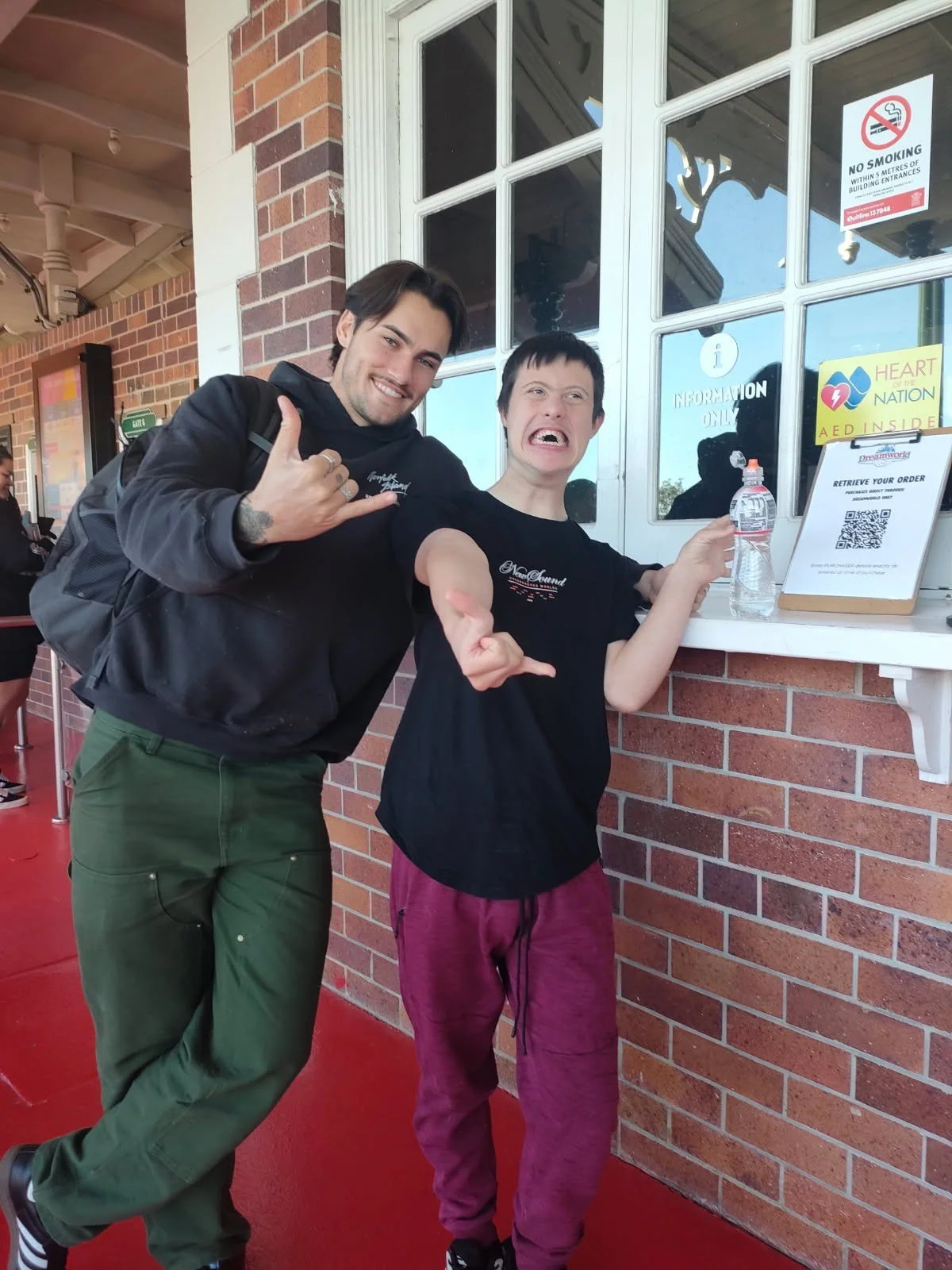 Matthew and Kristian standing beside a ticket window at Dreamworld. They are both smiling and displaying a cool sign with their right hands.