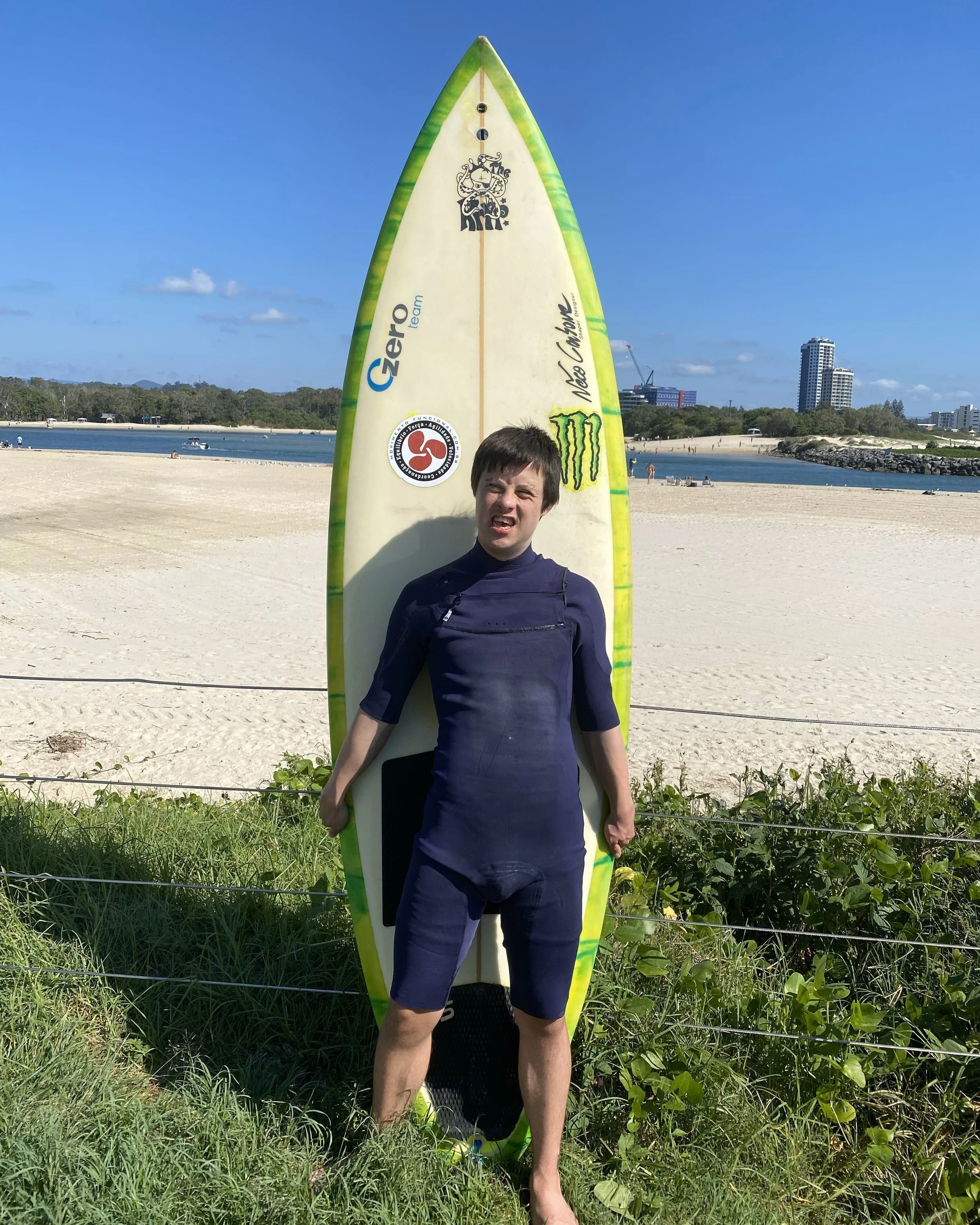 Photo of Matthew at the beach wearing a blue wetsuit. He is standing on the grass with a green and yellow surf board standing on it's end behind him. The sand and water are behind him.