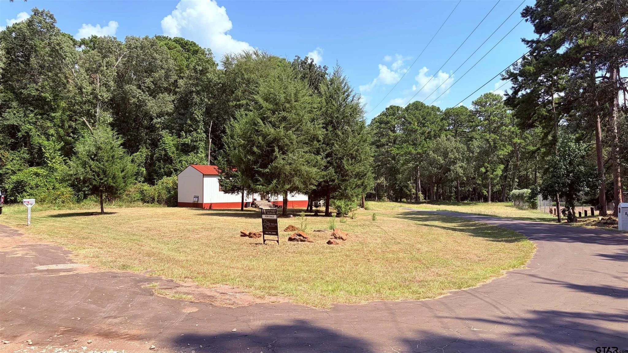 A small white building with a red roof is situated among tall green trees on a grassy lot. There is a 'For Sale' sign in front of the building, and a paved driveway curves around the lot. The sky is bright with some clouds, and there are power lines overhead.