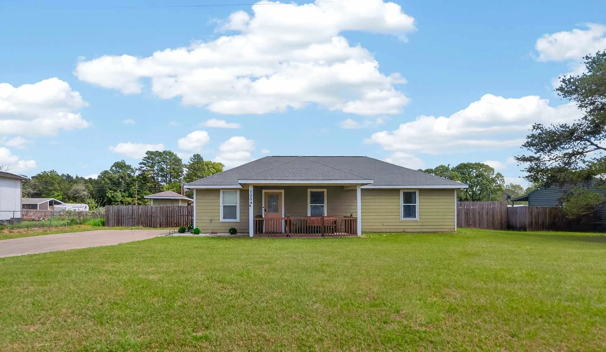 A yellow house with a front porch and a wooden railing, surrounded by a well-maintained lawn and a simple wooden fence, under a blue sky with white, fluffy clouds.
