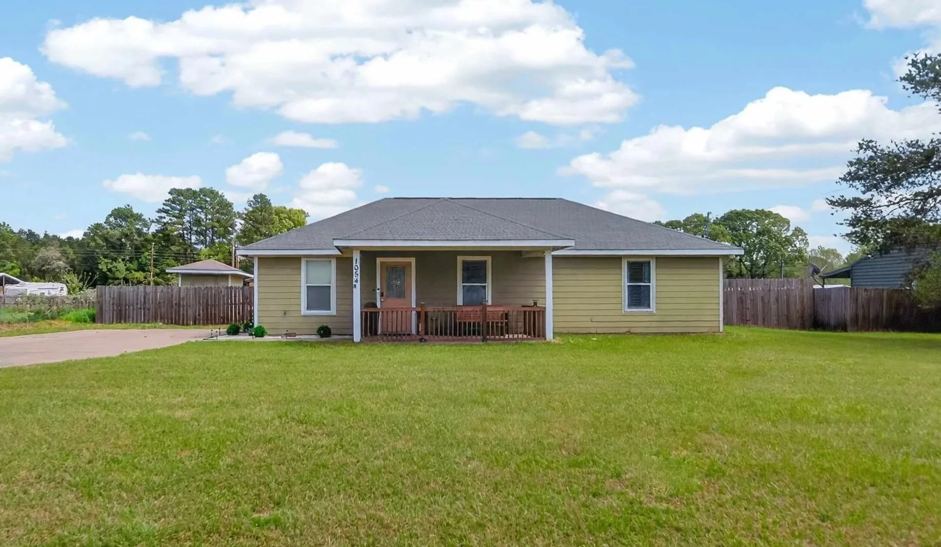 Single-story yellow house with a small front porch, surrounded by a green lawn and a wooden fence, under a partly cloudy sky.