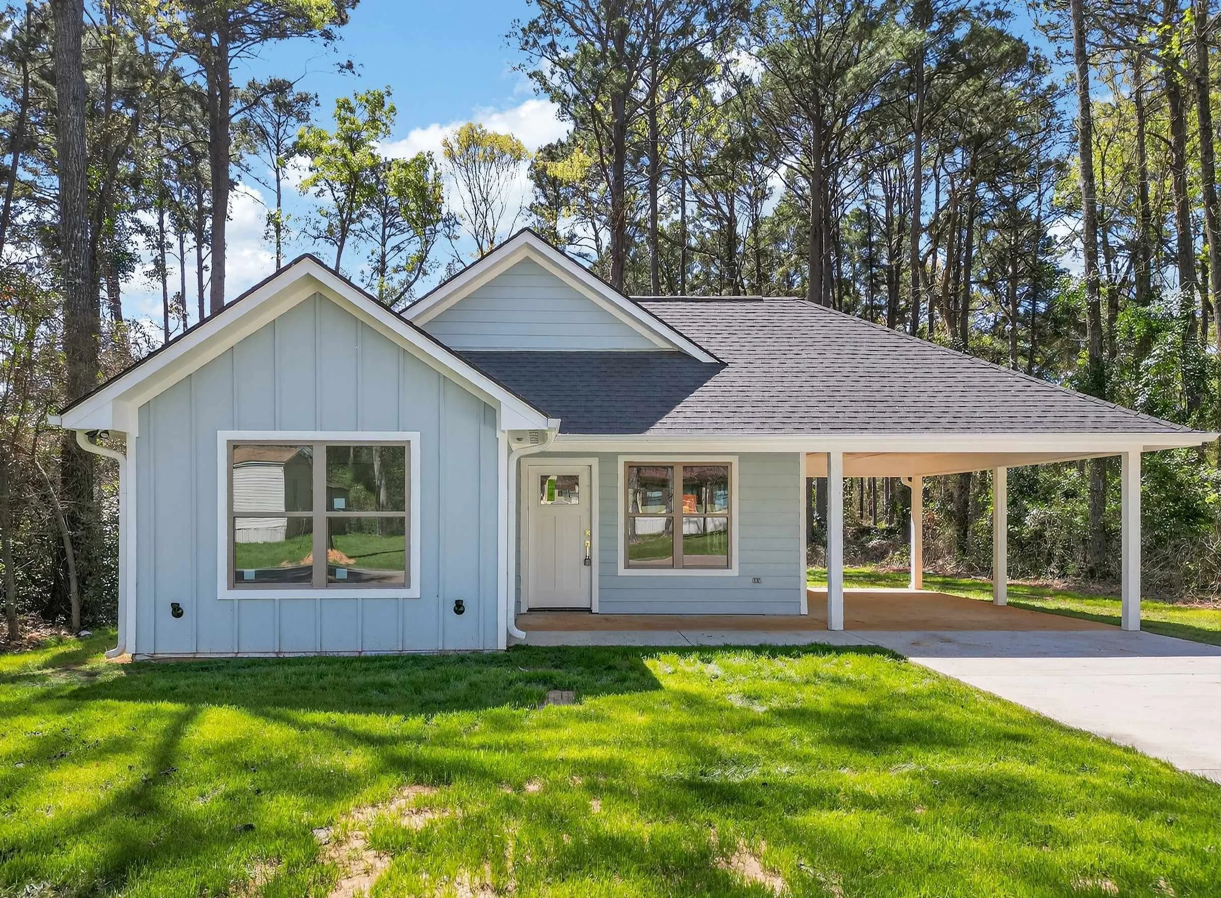 A modern single-story house with light gray siding, white trim, and a dark gray roof, surrounded by lush green grass and tall trees in the background on a sunny day.