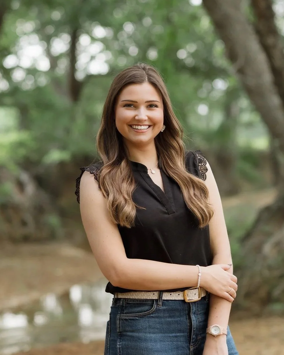 A young woman with long wavy brown hair smiling outdoors near a stream, wearing a black sleeveless blouse, jeans, and jewelry.