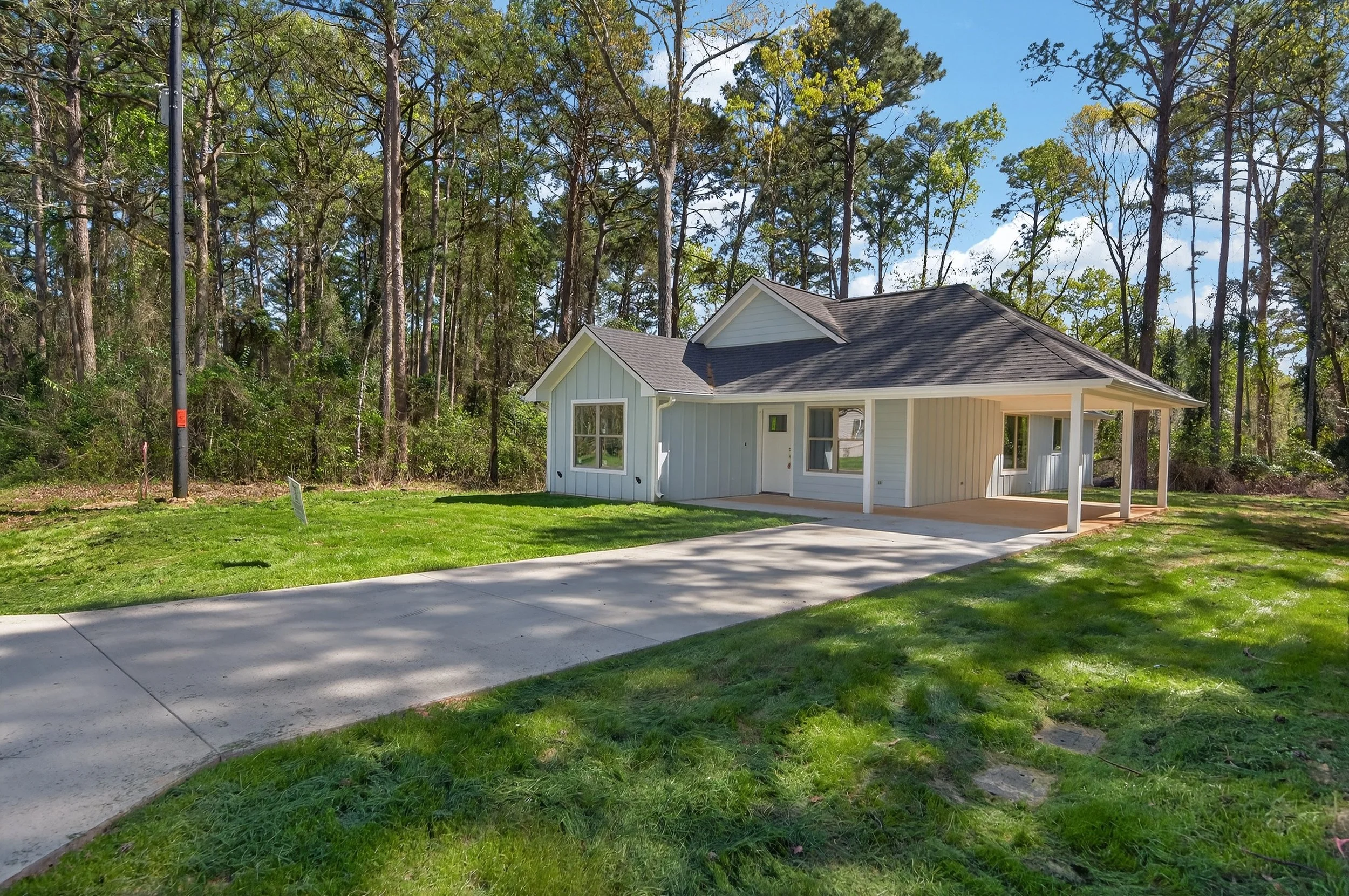 A small white house with a porch on a grassy lawn, surrounded by tall trees with green leaves, under a bright blue sky with clouds.