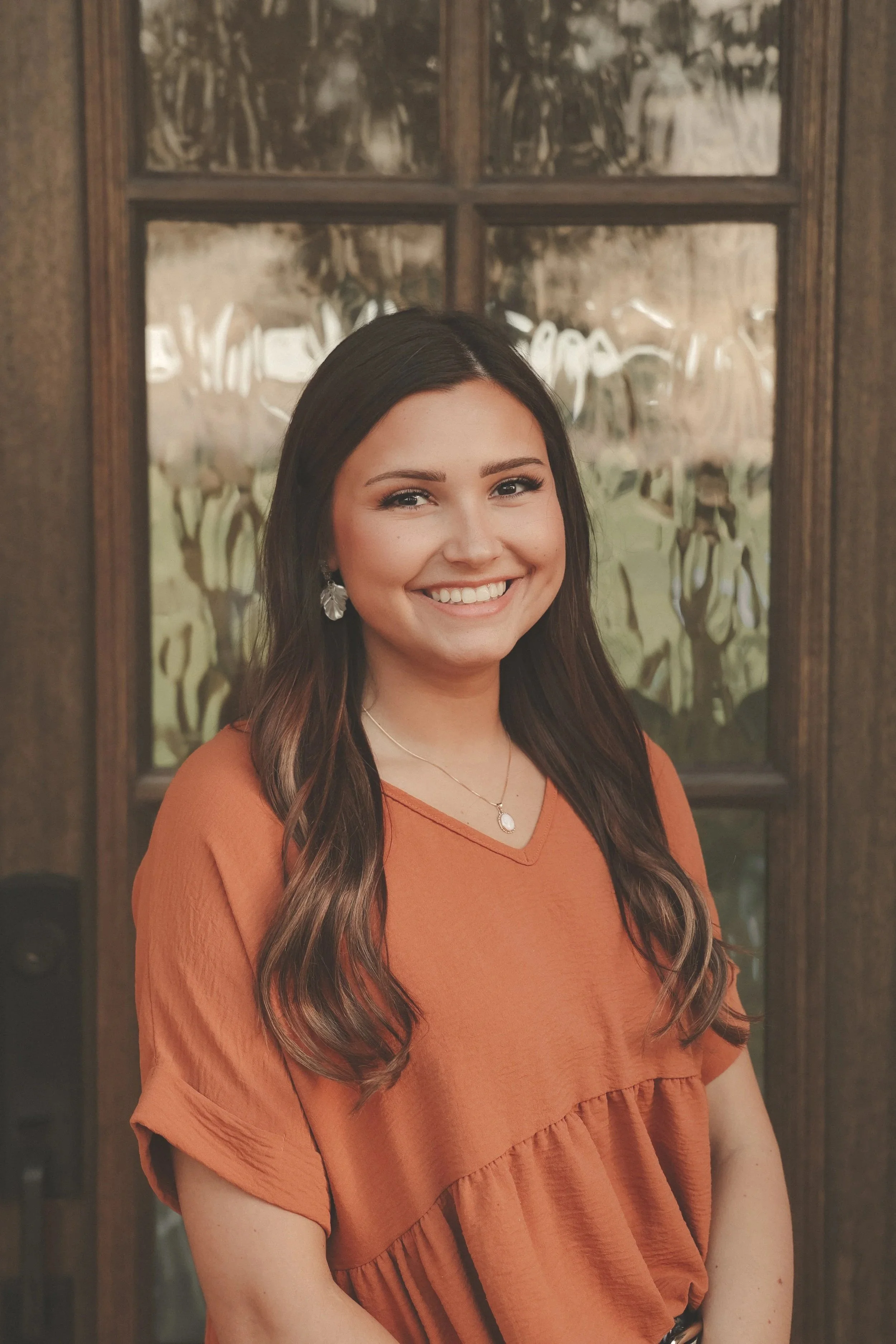 A woman with dark brown hair and earrings smiling in front of a window with a wooden frame.