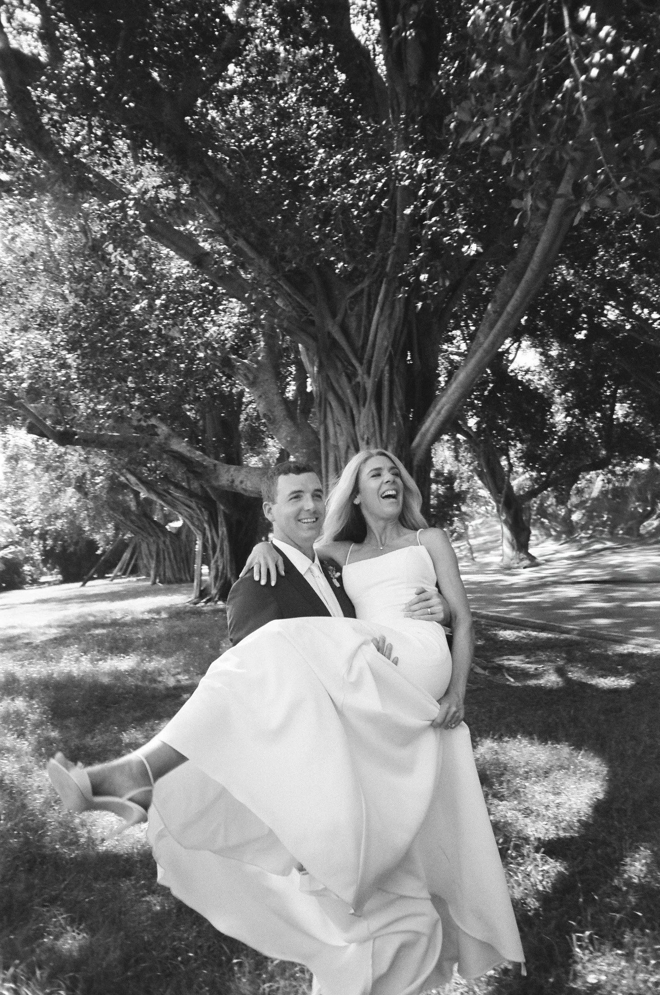 A black and white photo of a bride and groom smiling while walking outside under a large tree, with the groom carrying the bride.