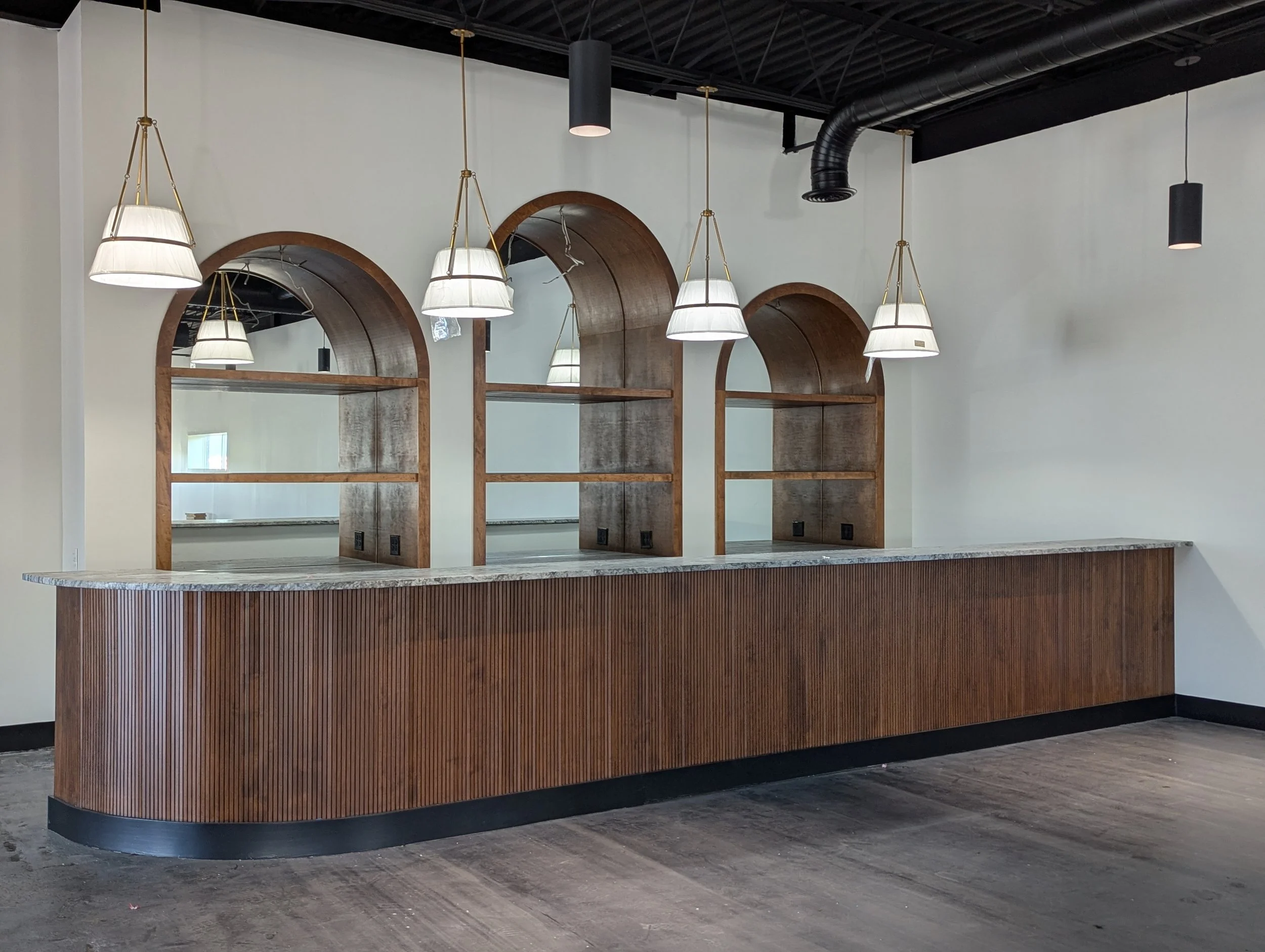 Modern kitchen with white countertops, dark gray and white cabinets, dark wooden floor, stainless steel refrigerator, black pendant lights, and large windows.