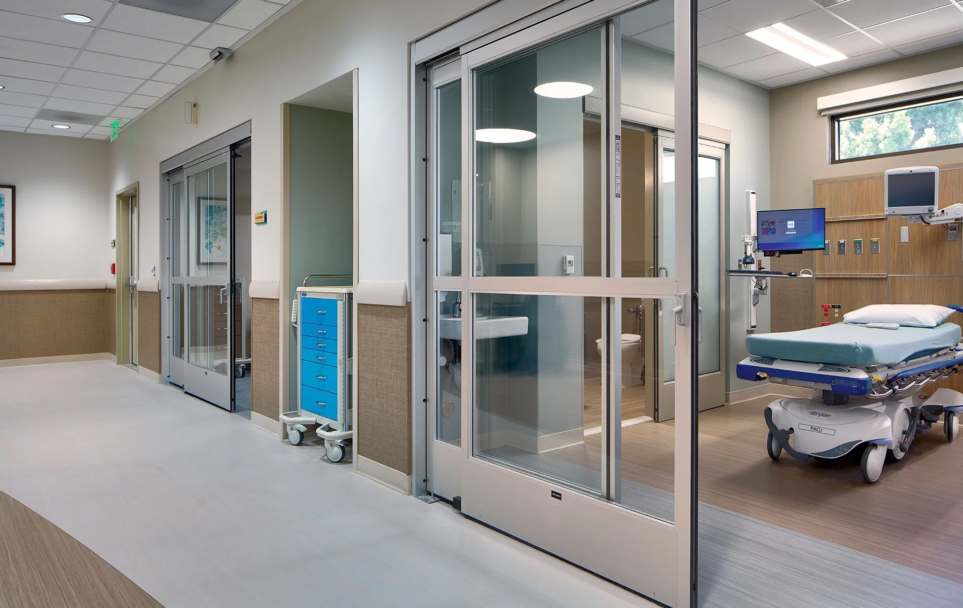 Empty hospital room with a hospital bed, medical equipment, and a glass-enclosed bathroom in the background.