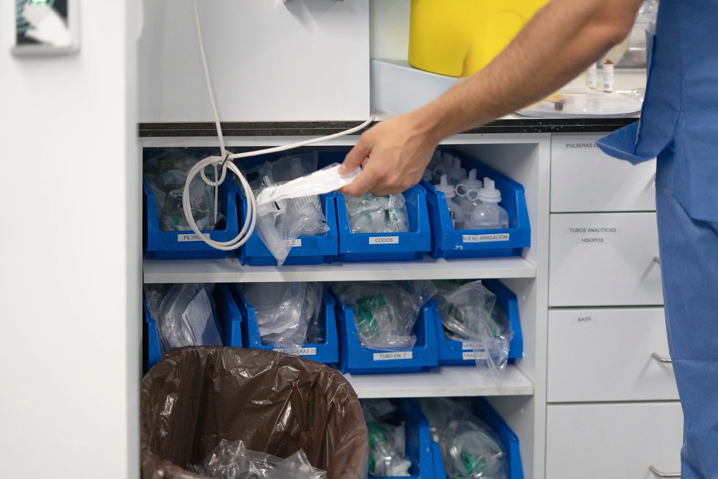 A healthcare professional's hand reaching into a medical supply cabinet with blue labeled bins containing medical supplies and equipment.