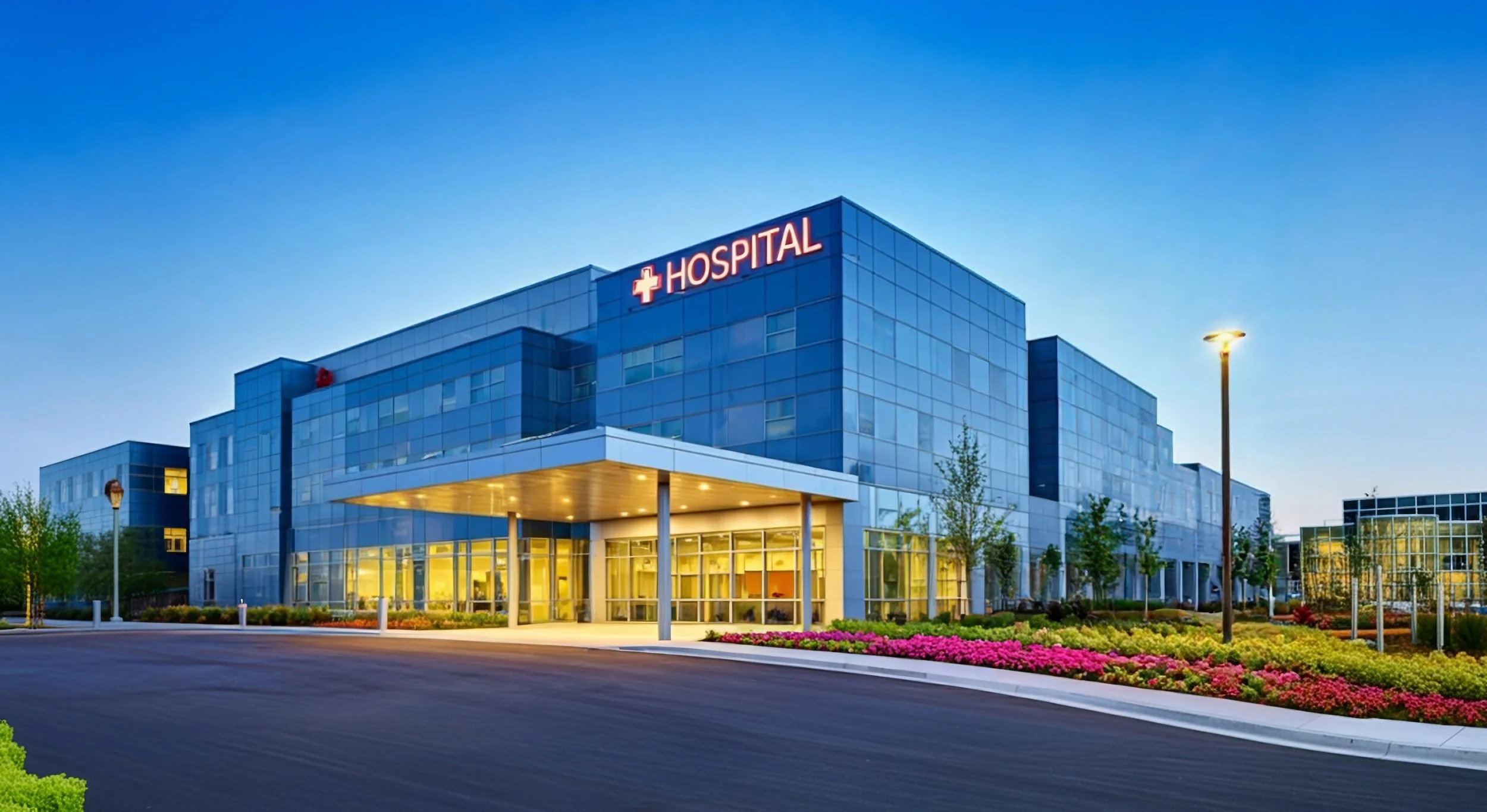 Modern hospital building with a glass facade, illuminated entrance, and a red medical cross sign with the word 'HOSPITAL' on top, surrounded by landscaped gardens and streetlights at dusk.