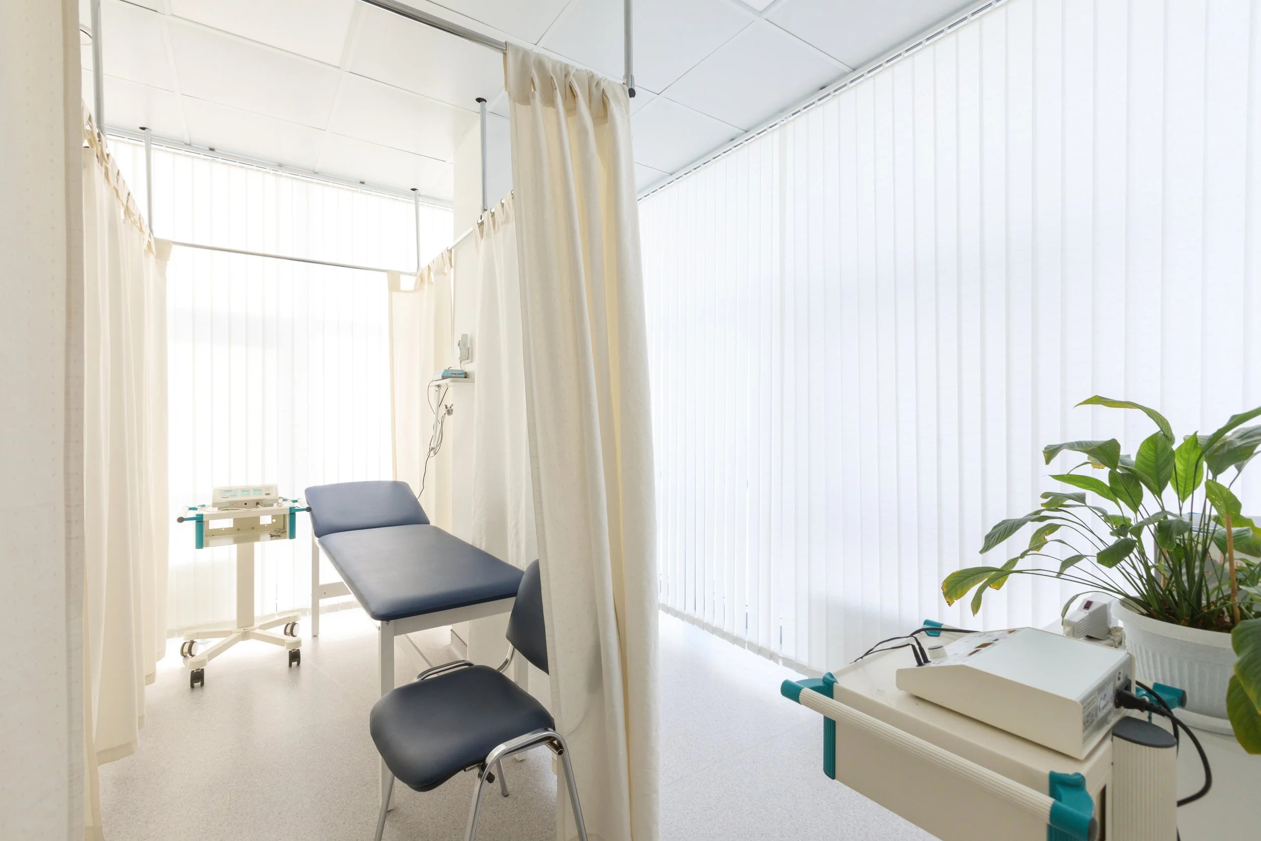 Empty hospital room with examination bed, privacy curtain, medical equipment, and a plant.