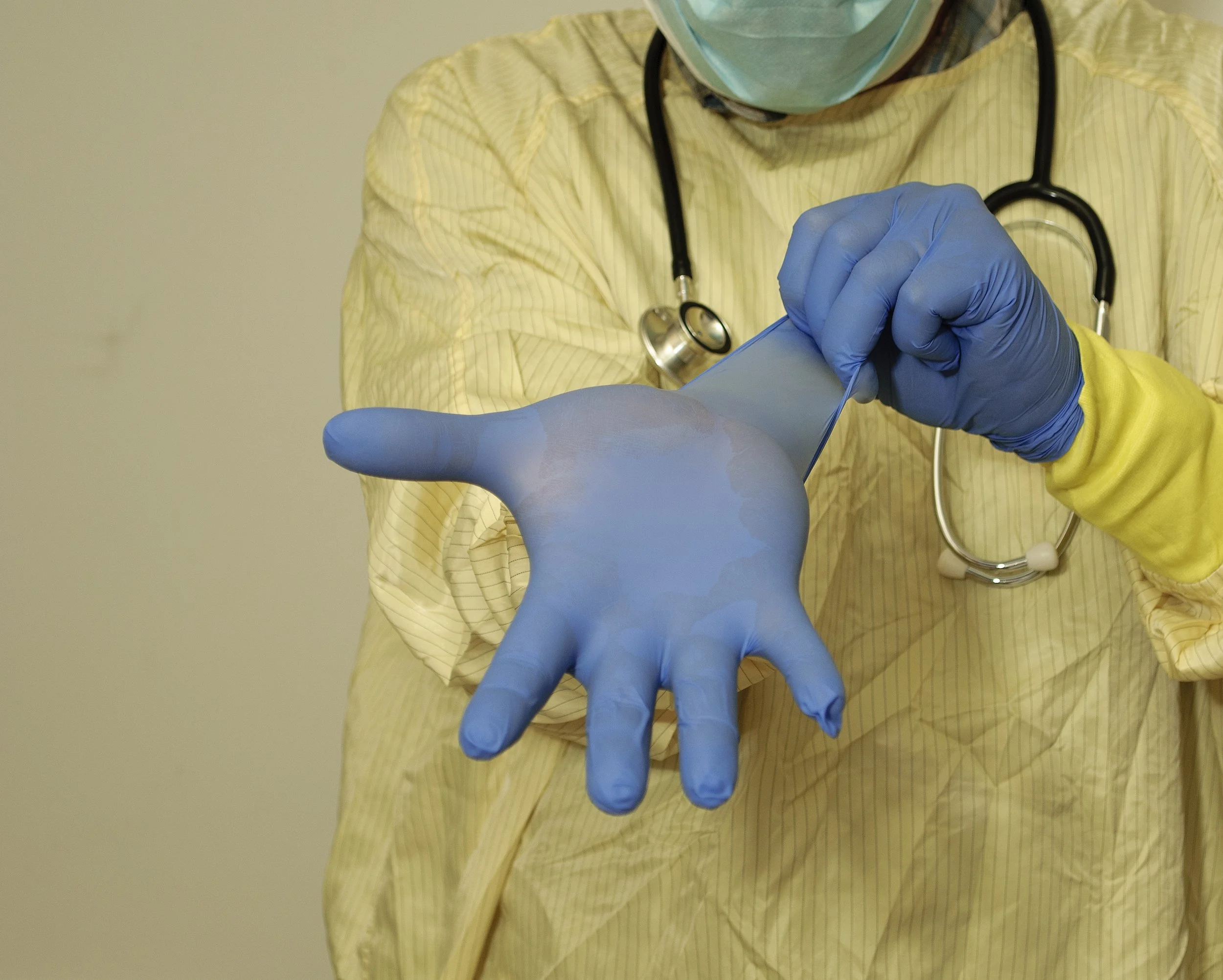 Medical professional in a yellow gown and blue gloves using a spatula to handle a large drop of liquid or gel, with a stethoscope around their neck.
