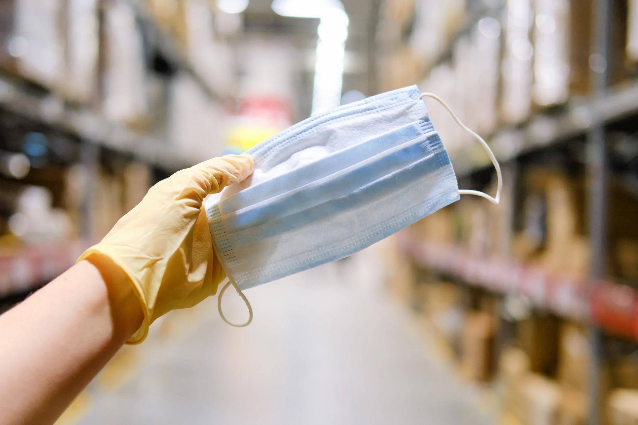 Person wearing a yellow glove holding a blue face mask in a warehouse aisle.
