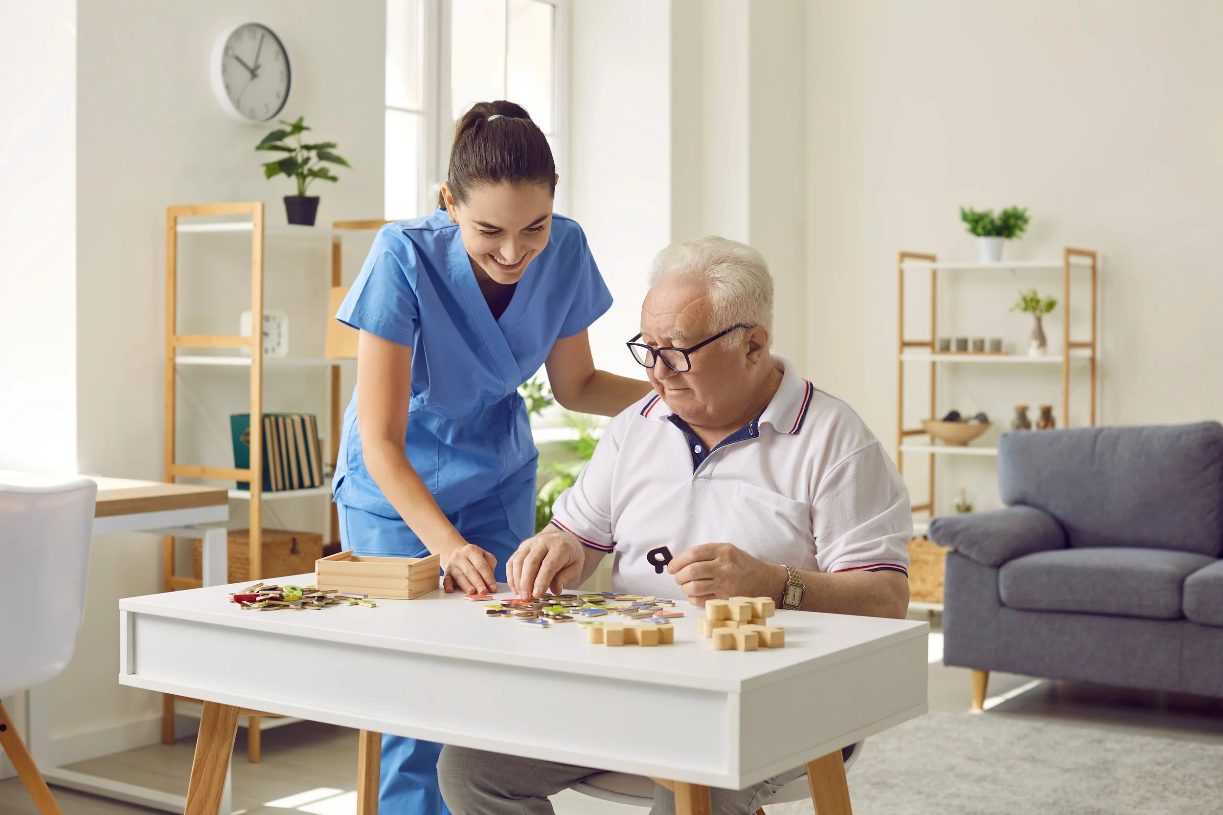 A caregiver and an elderly man playing a board game together at a white table in a bright, modern living room.