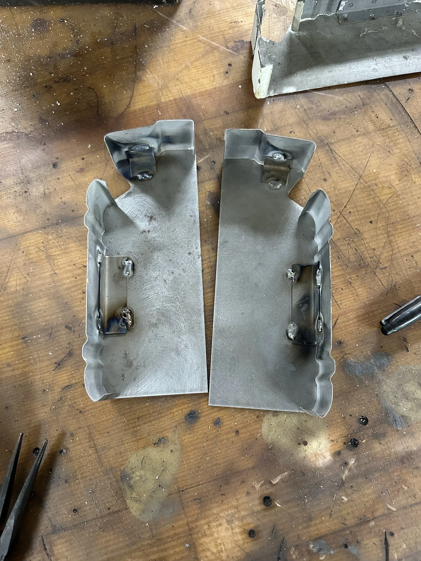 Two metal brackets with welded mounting points on a wooden workbench.