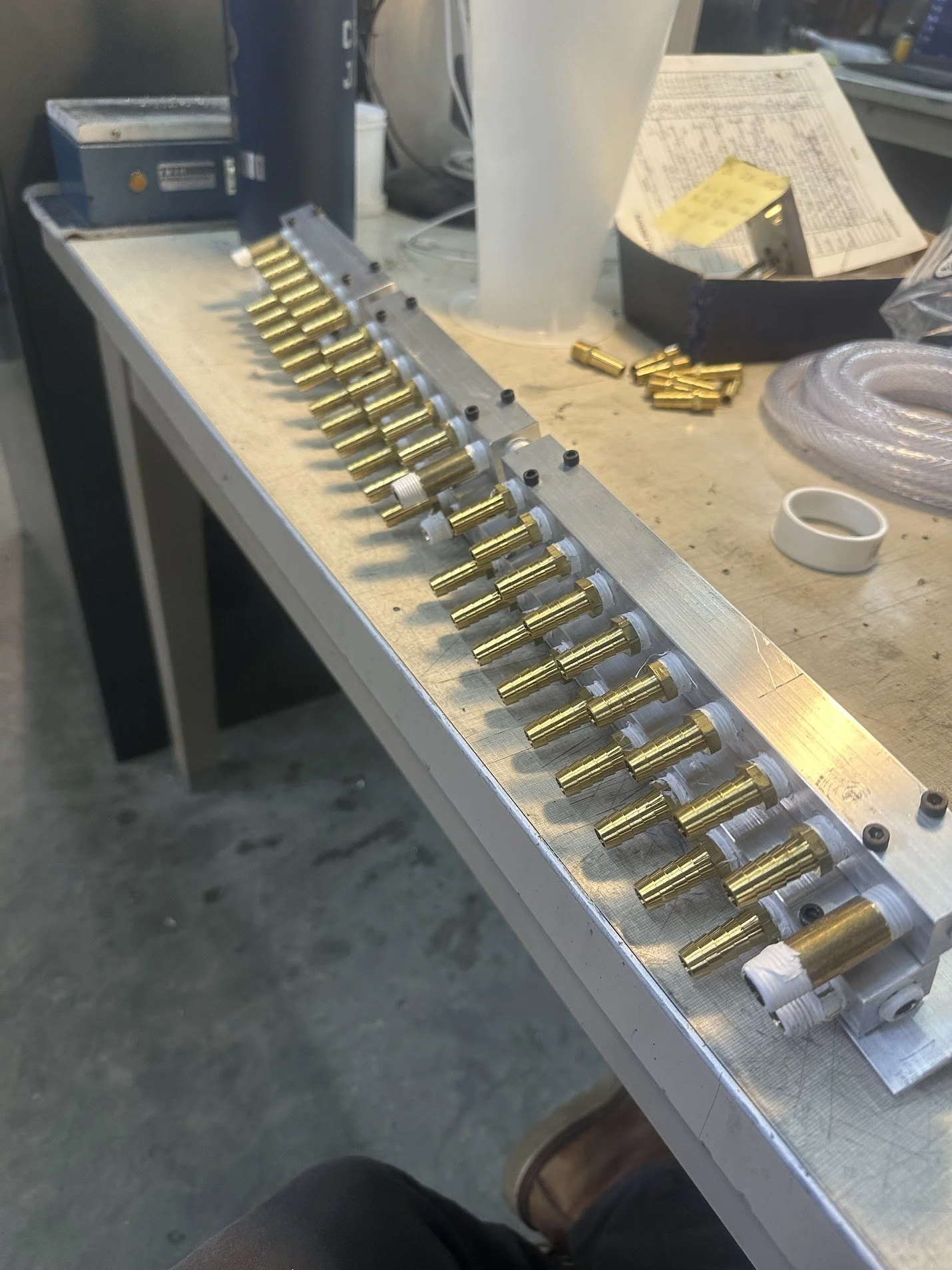 A workbench with multiple gold-colored metallic components aligned in a row, along with small black parts, a roll of white tape, coiled tubing, and various tools and papers nearby.
