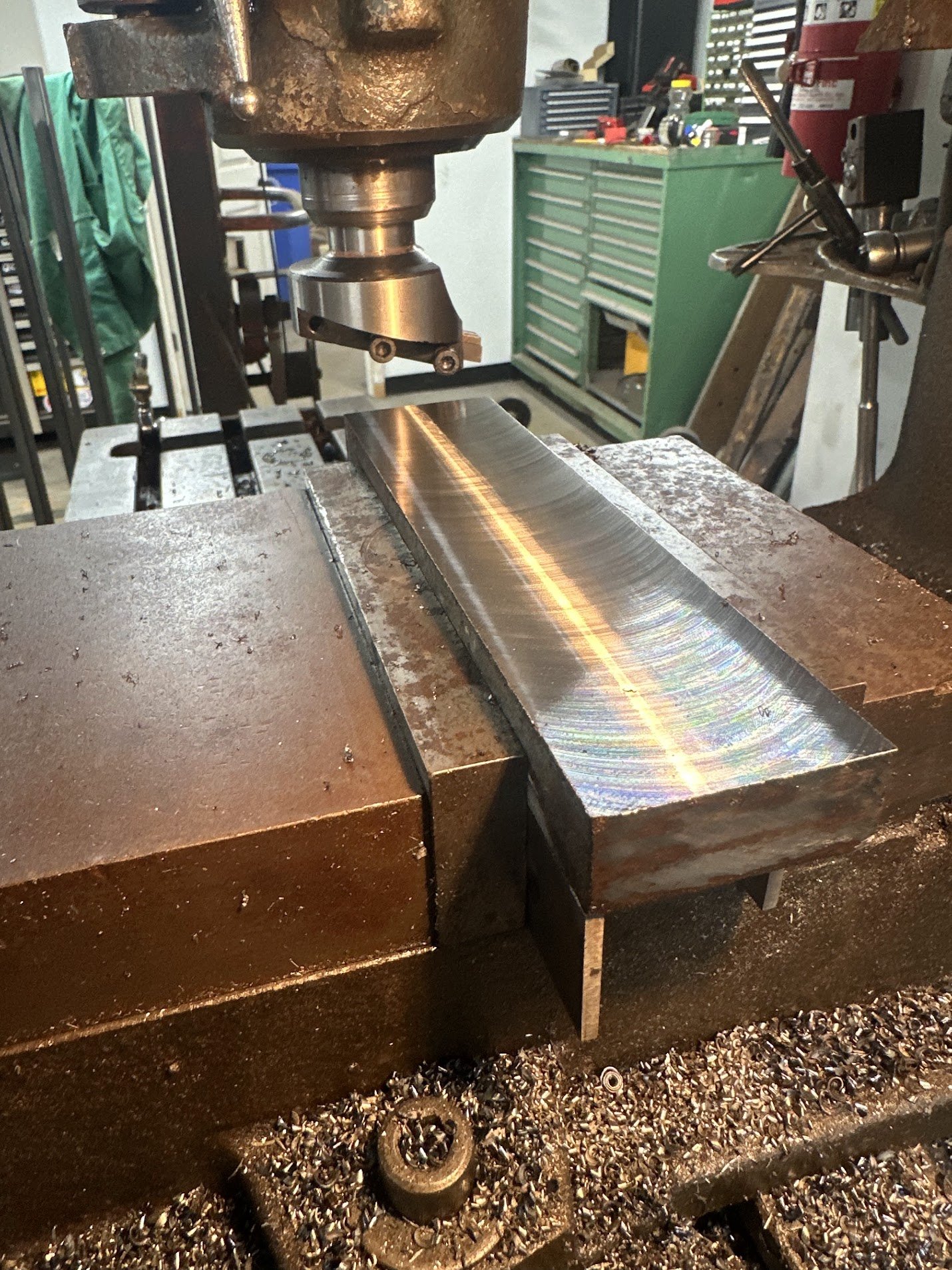 Close-up of a metalworking machine shaping a metal piece, with a workbench and toolbox in the background.