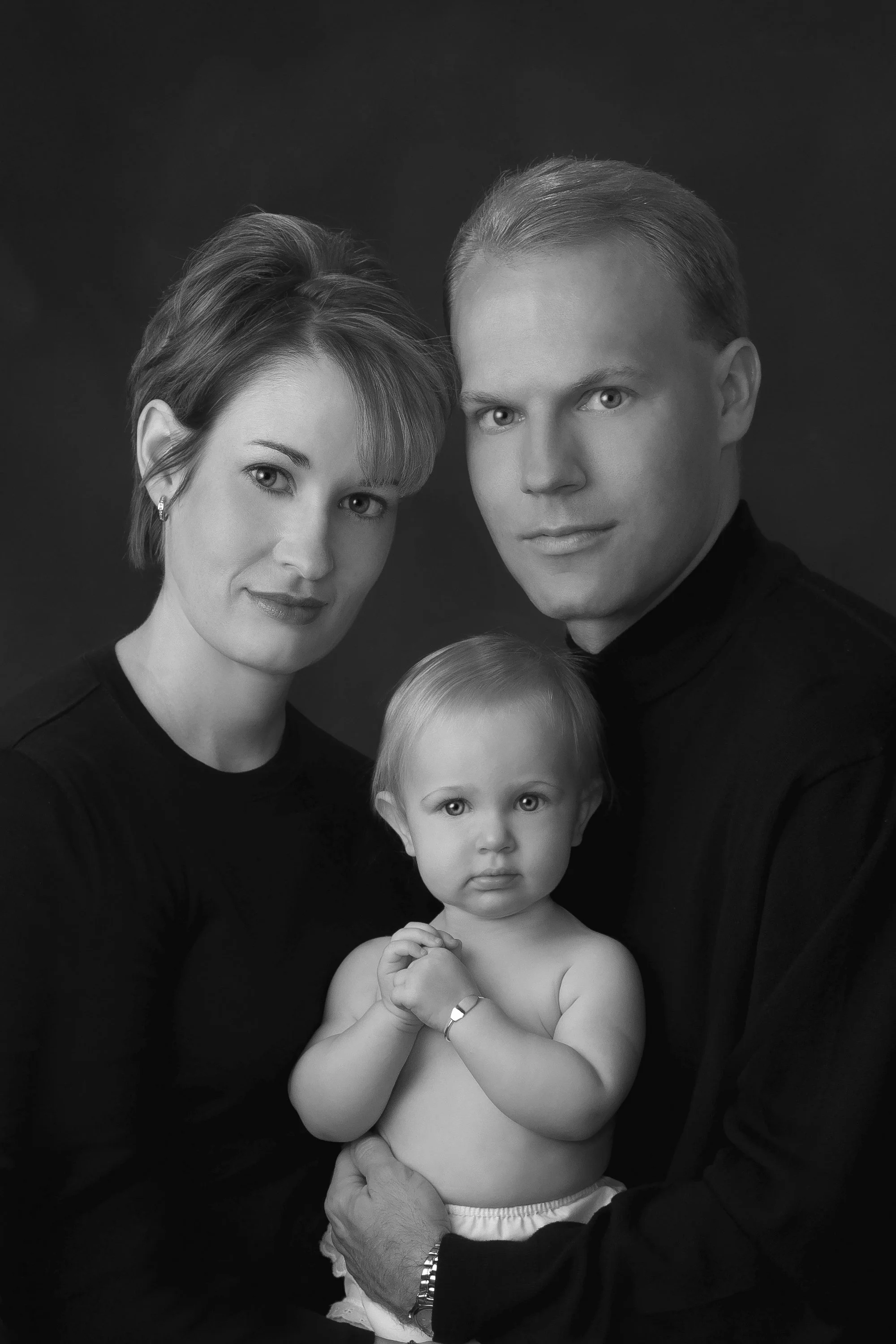 Black-and-white studio portrait of a mother and father with their baby girl, posed together against a dark background.
