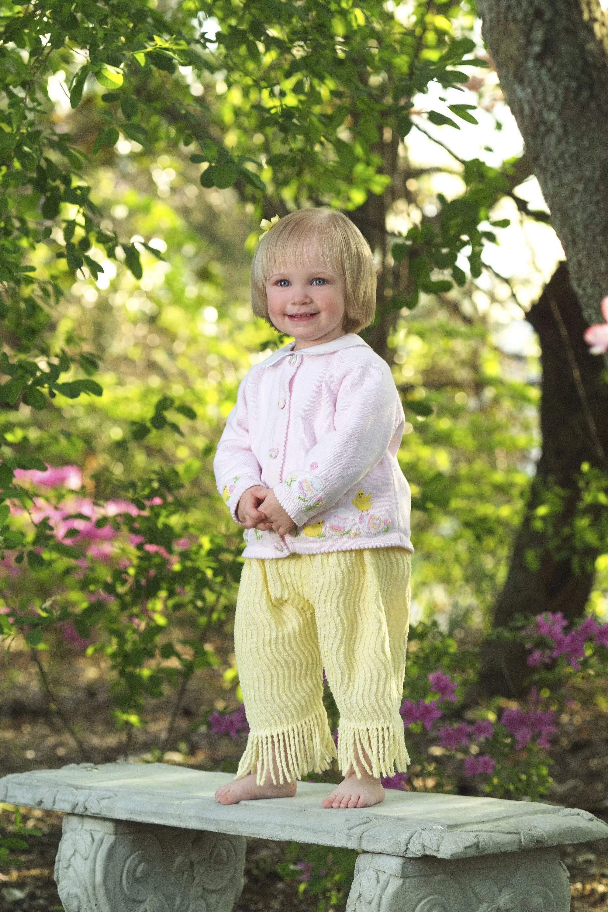 Blonde toddler girl wearing a pink cardigan and yellow pants, standing on a stone garden bench surrounded by greenery.