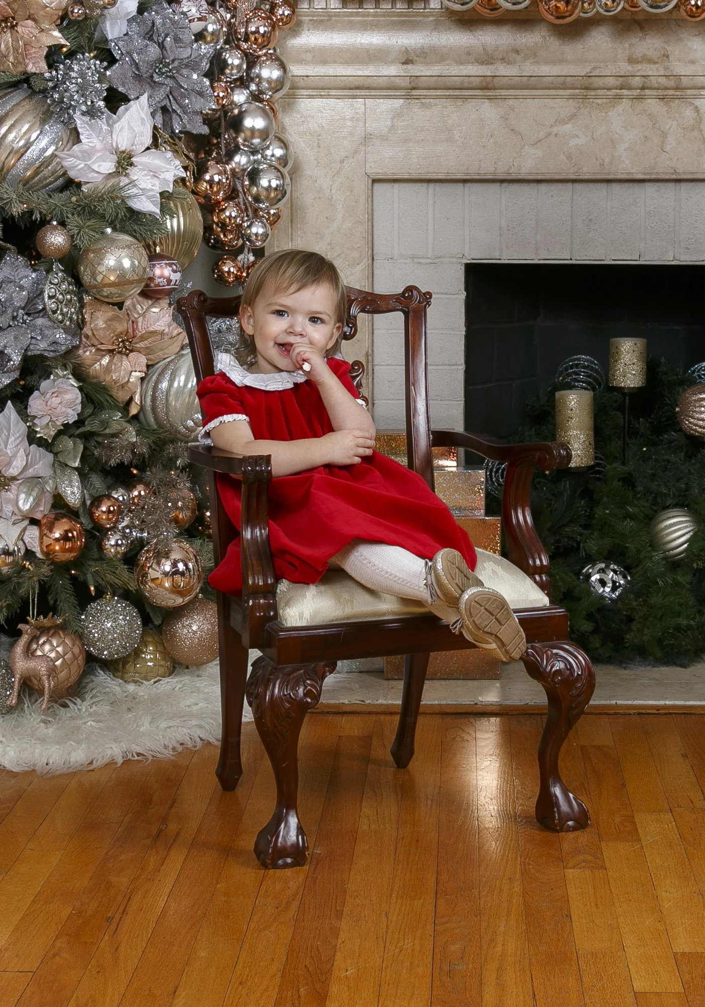 Young girl in a red holiday dress seated in a wooden chair at Hayes & Fisk’s Richmond studio, with a decorated Christmas tree and fireplace behind her.
