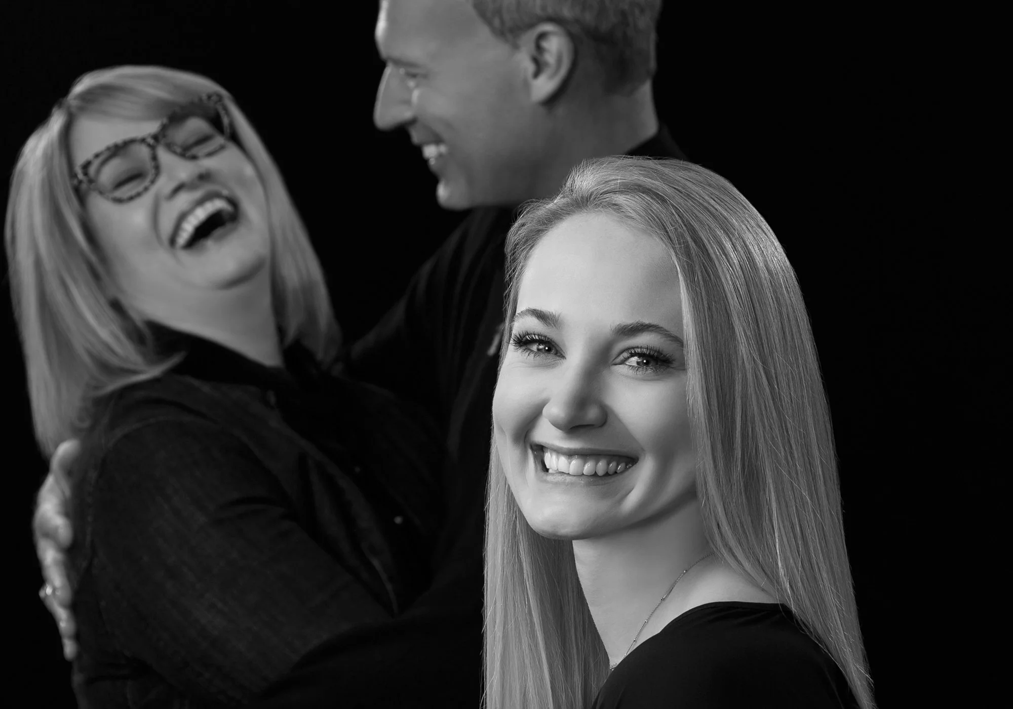 Black-and-white family portrait with a smiling young adult daughter in the foreground and her mother and father together behind her.